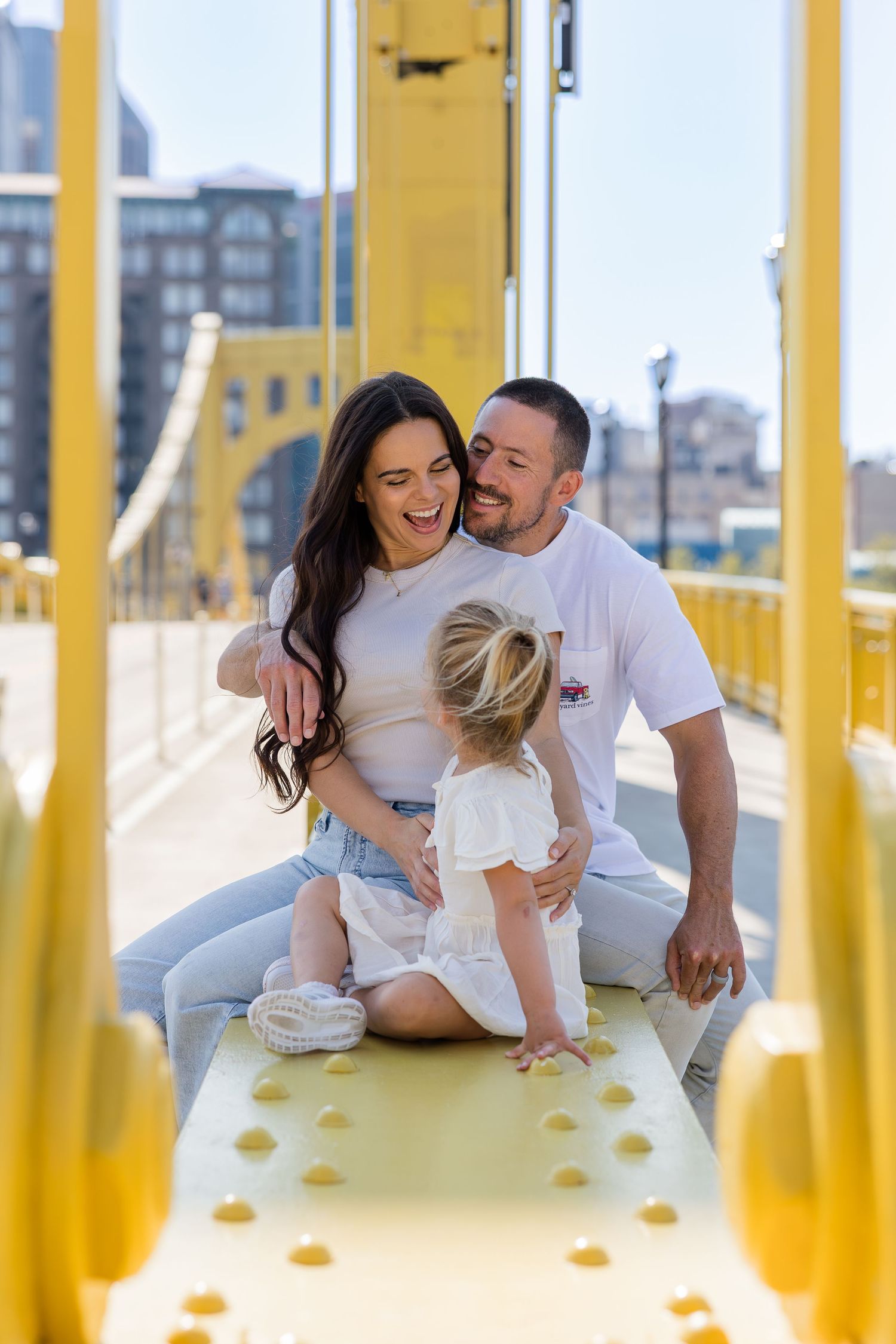 Smiling couple sits with their toddler daughter on a bright yellow steel bridge with city buildings in the background.