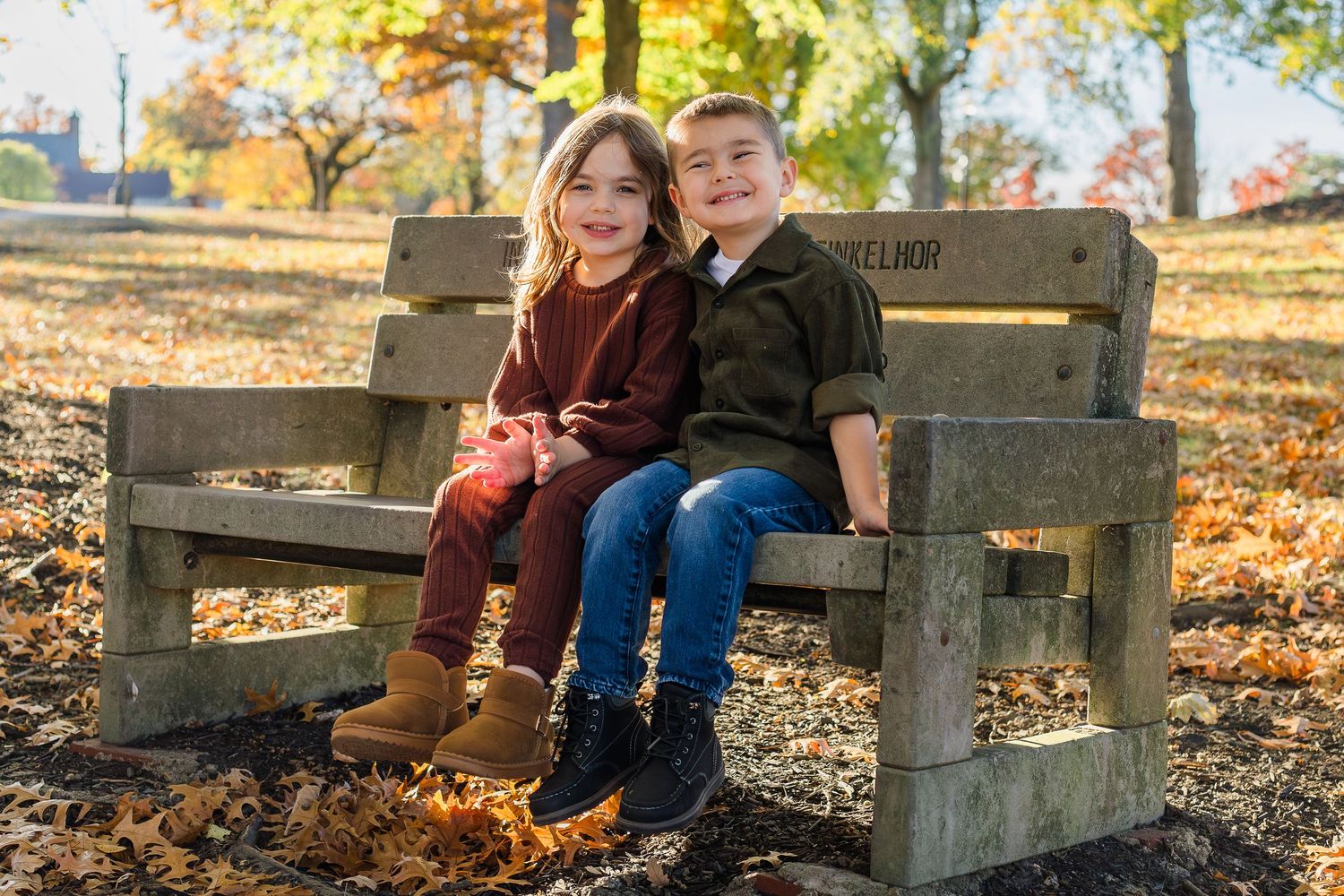 Two smiling children sit side by side on a rustic wooden bench surrounded by fallen leaves in a sunny autumn park.