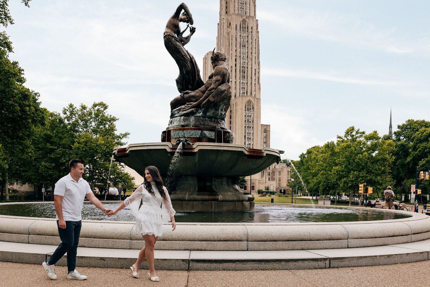 Couple holding hands and walking beside a sculpted stone fountain with a tall Gothic cathedral tower rising behind green trees.