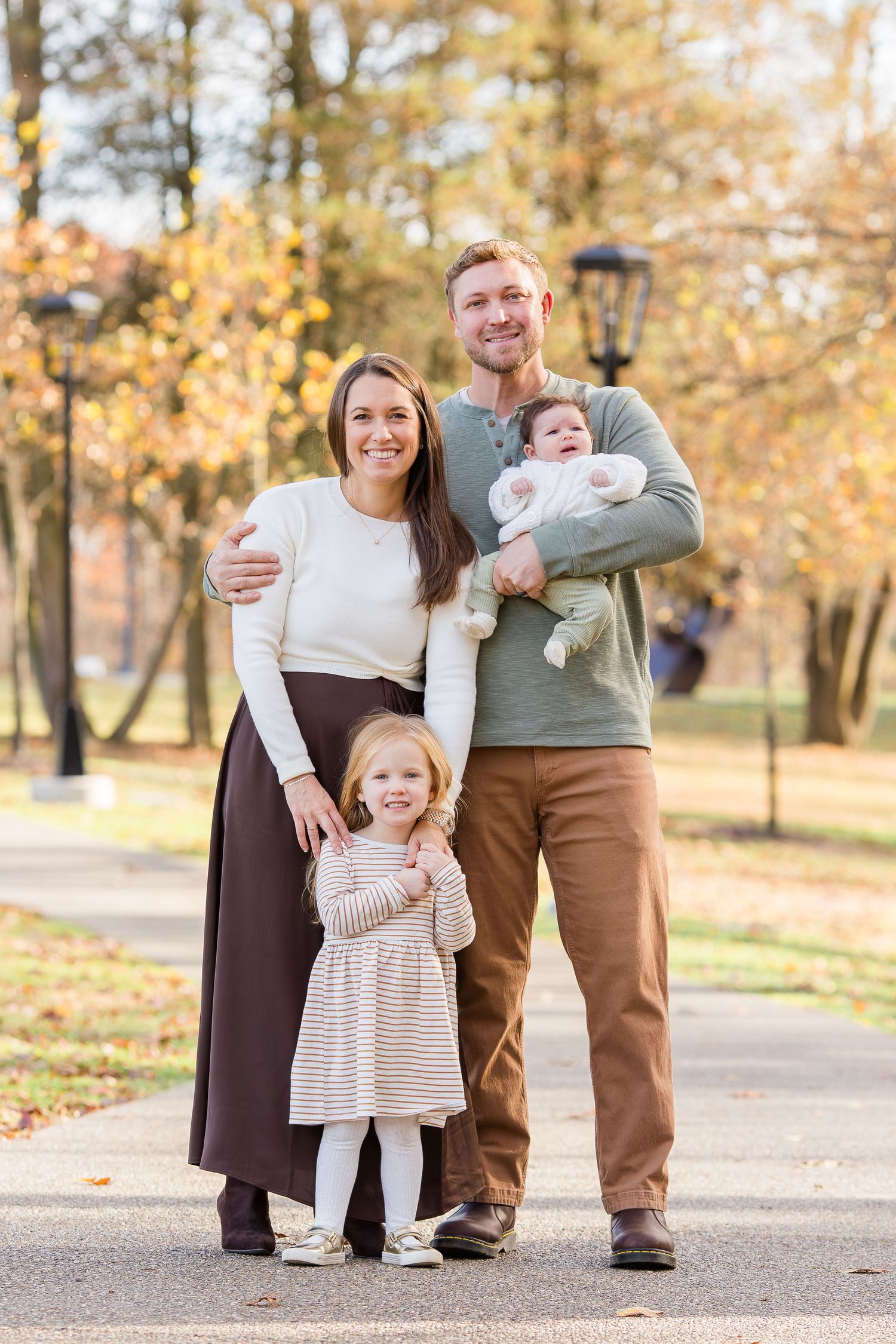 Smiling family of four with a baby and toddler girl posing on a park path surrounded by golden autumn trees.