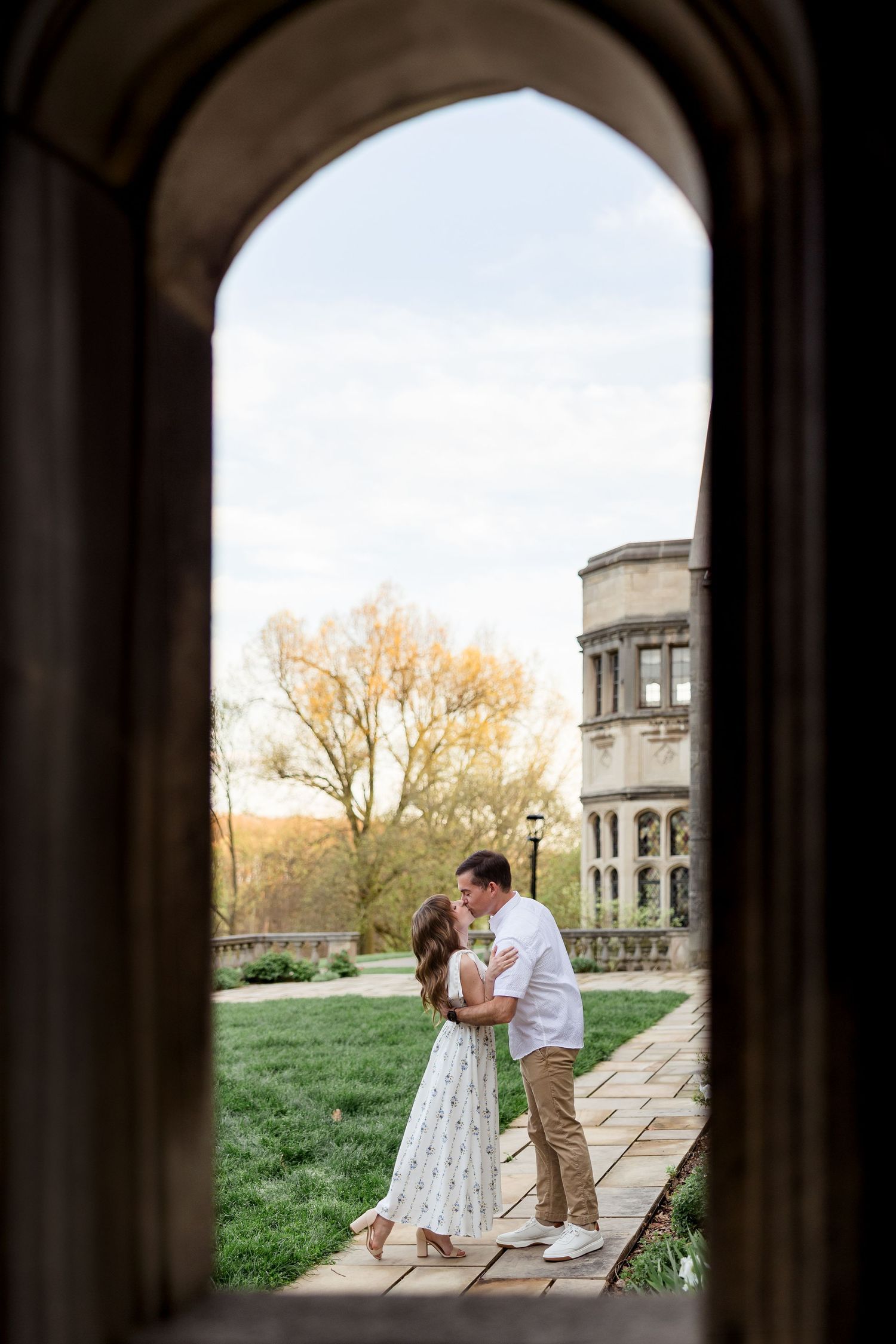 Couple kissing on a stone garden path, framed by a gothic arched window with a historic castle and spring trees in the background.