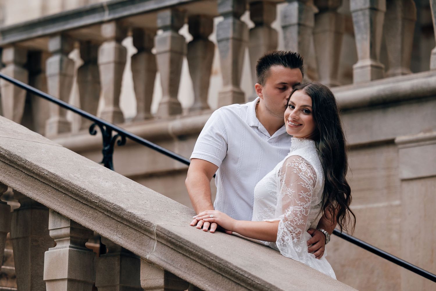 A man in a white polo kisses a smiling woman in a white lace top on the cheek as they pose on a stone staircase with balustrades.