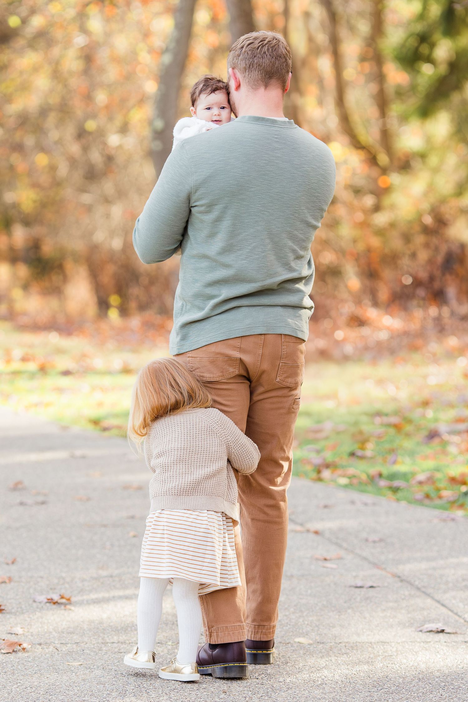 A father in a green sweater holds a baby while a toddler hugs his leg on a path surrounded by colorful autumn trees.