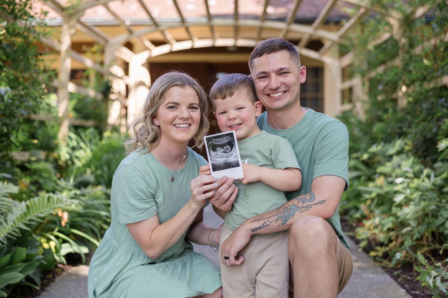 Smiling couple and their toddler son in matching green outfits holding an ultrasound photo in a sunny garden, announcing a pregnancy.