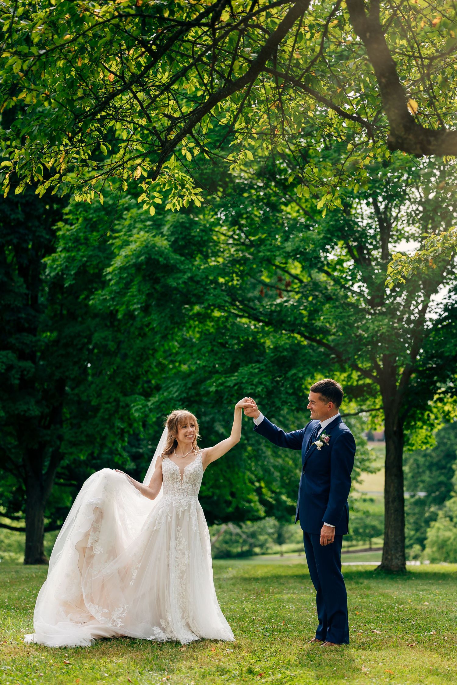 Bride in a flowing lace wedding gown twirls while holding hands with the groom in a navy suit on a sunny, tree-lined park lawn.