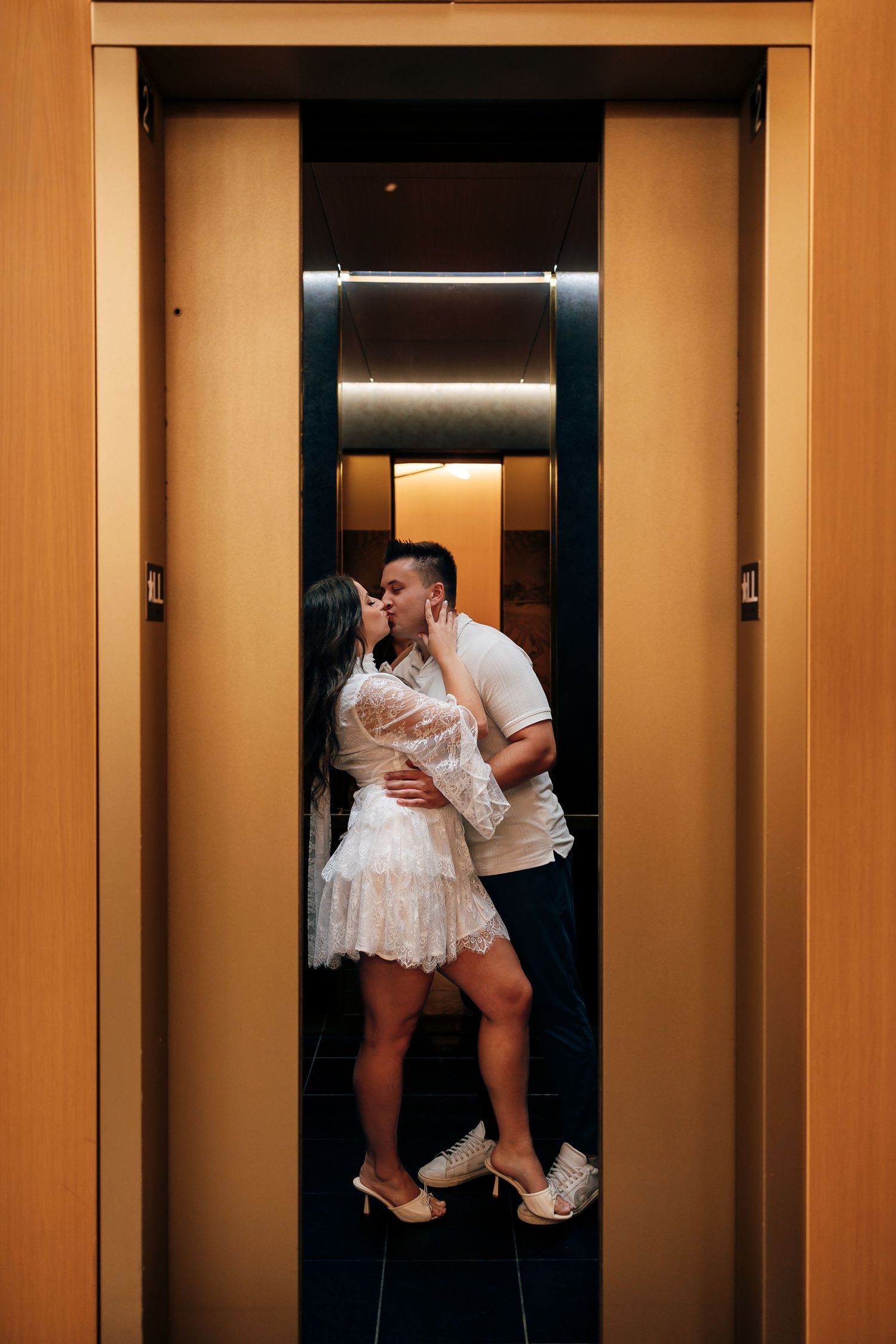 Couple kissing inside a wood-paneled elevator, the woman in a white lace dress and heels, the man in a white t-shirt and sneakers.