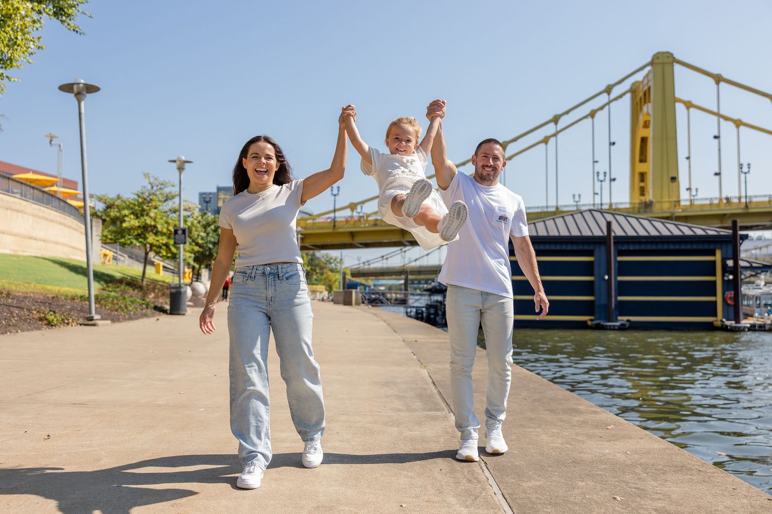 Smiling couple holding hands and swinging a toddler between them while walking along a sunny riverside promenade near a yellow steel bridge.