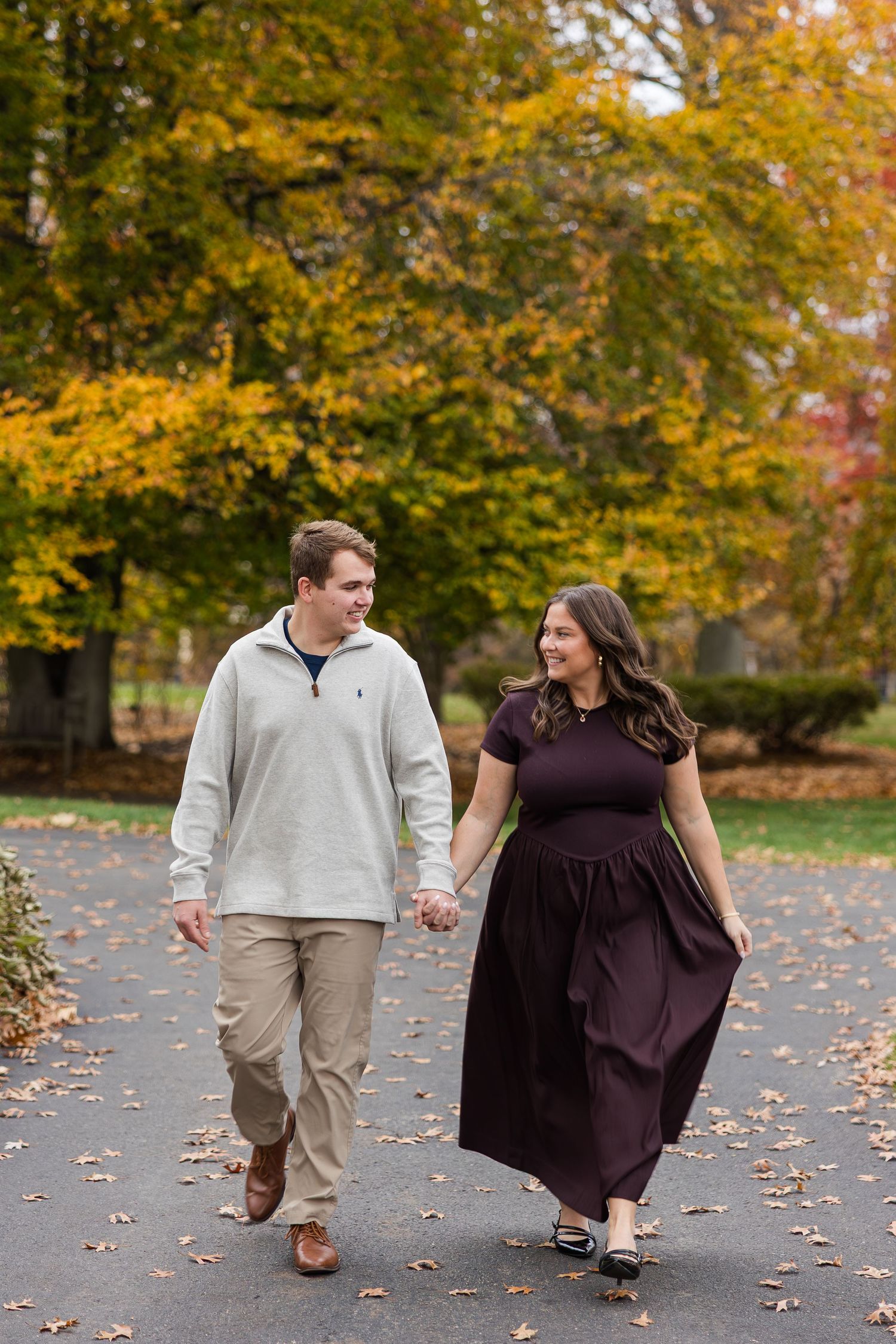Smiling couple holding hands while walking along a leaf-covered path beneath colorful fall trees during an autumn engagement photo session.