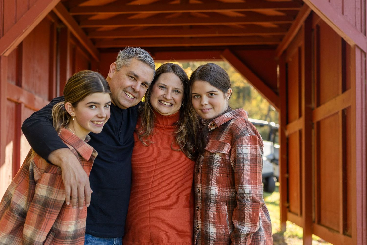 A smiling family of four poses closely together inside a rustic red barn, wearing plaid shirts and an orange sweater on a sunny autumn day.