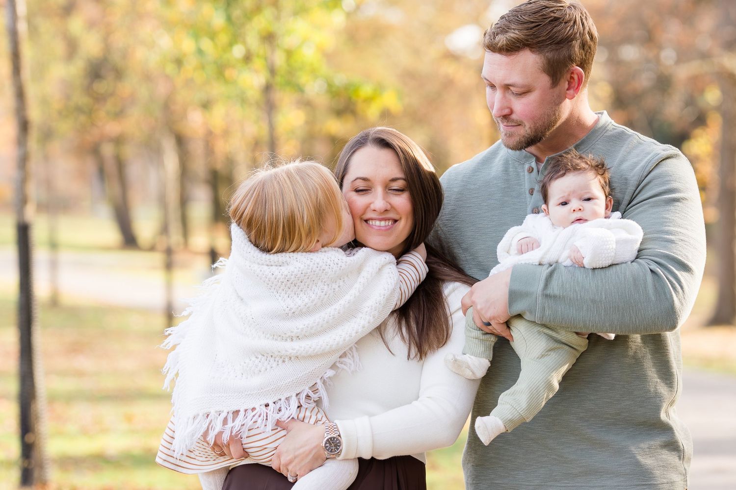 Smiling family of four embracing outdoors in a sunlit autumn park, with a mother and father holding a toddler and a baby.