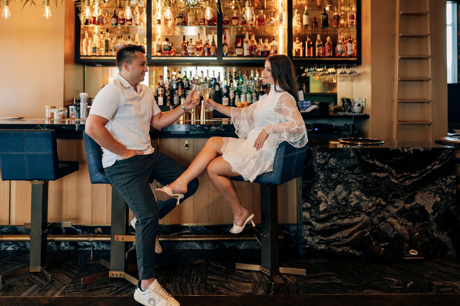 Couple toasting cocktails while seated on bar stools at an elegant bar with a backlit liquor shelf and marble counter.
