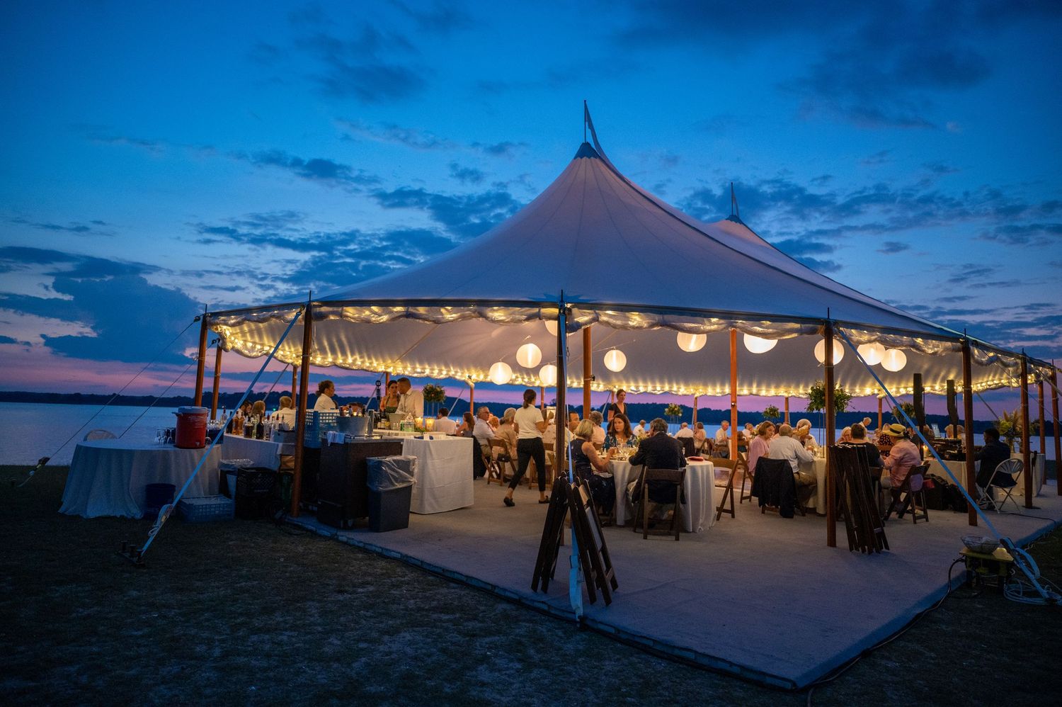 Illuminated white party tents glow against a dusky blue sky during an outdoor evening event at sunset.