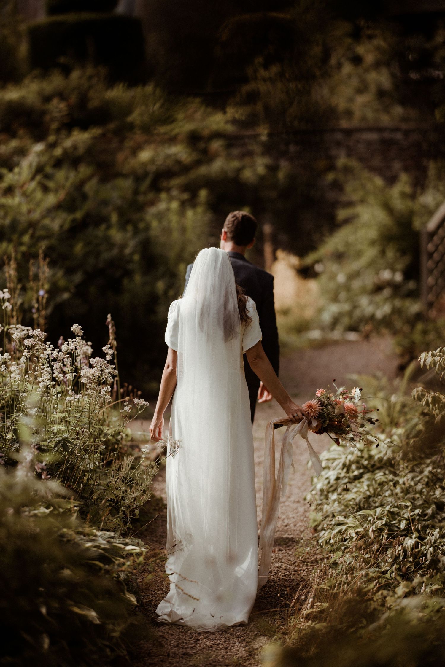 A man in a wedding suit & a woman in a flowing white wedding dress walking through a wildflower garden at Askham Hall.
