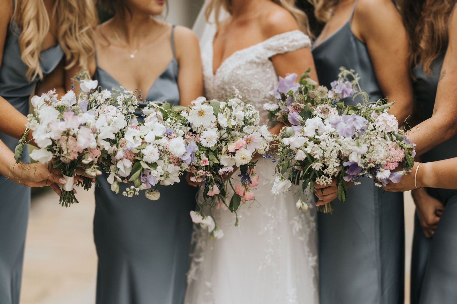 Wedding party holds elegant bouquets of white and purple flowers against soft grey bridesmaid dresses and white bridal gown.