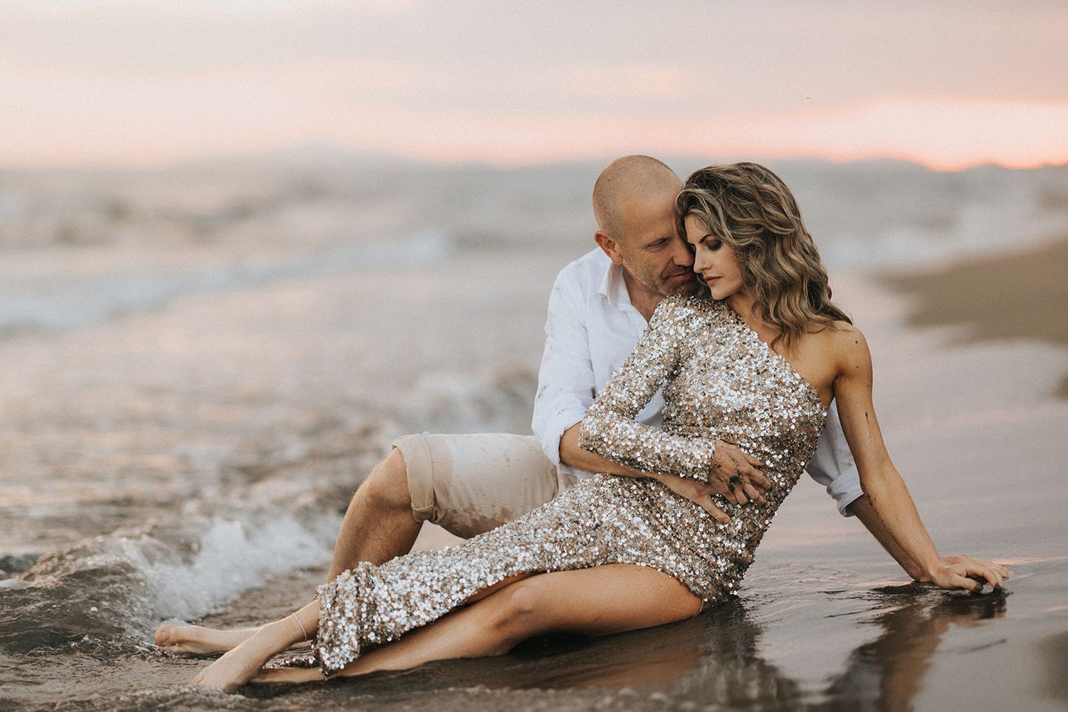 Couple in elegant attire poses intimately on international beach during golden hour sunset.
