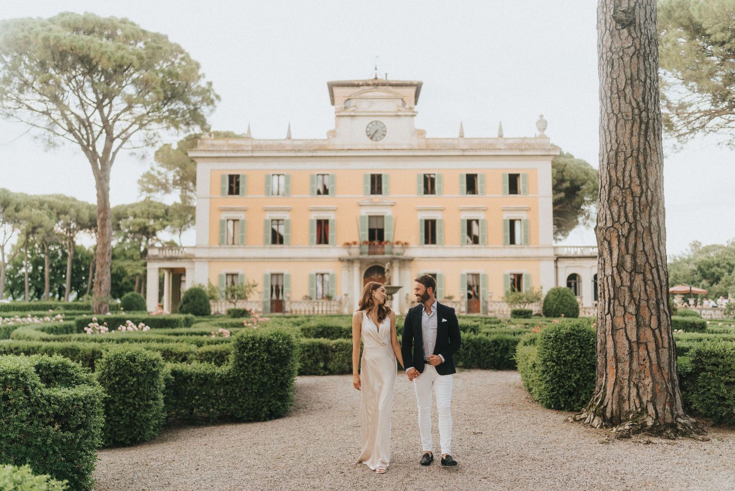 Elegant couple stands on a gravel path in front of a beautiful Italian villa with manicured hedges and tall pine trees.