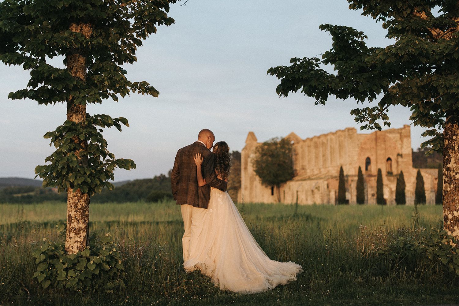 Two figures in wedding attire stand close together at sunset near an ancient stone building with cypress trees.