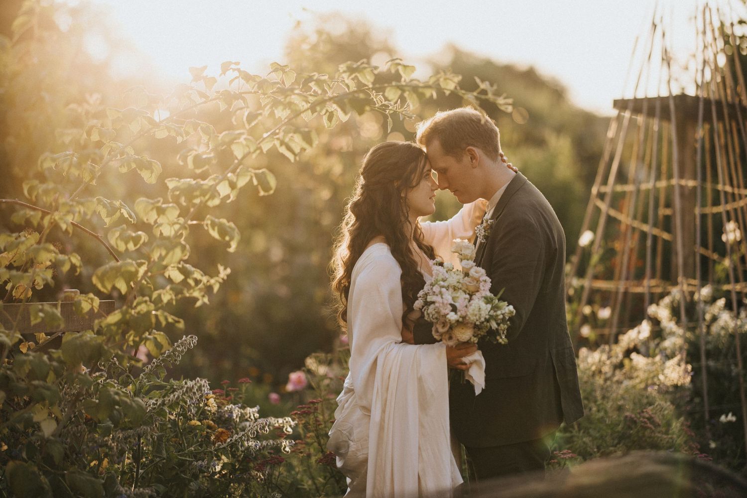 A couple shares an intimate moment in a sunlit garden with roses and a floral bouquet at golden hour at Askham Hall.