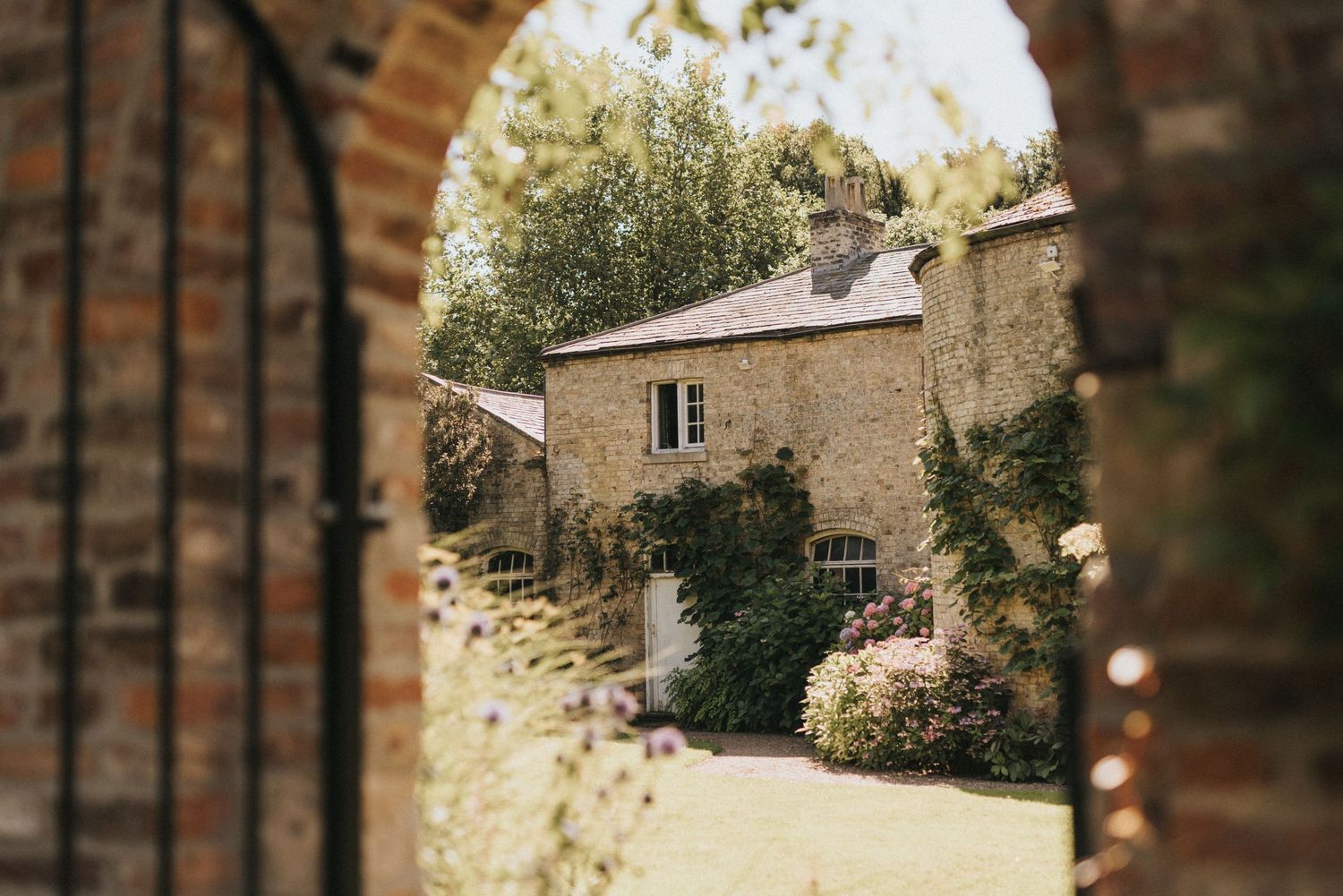 View through a brick archway of a charming stone cottage at Saltmarshe Hall with climbing vines and garden plantings in soft natural light.