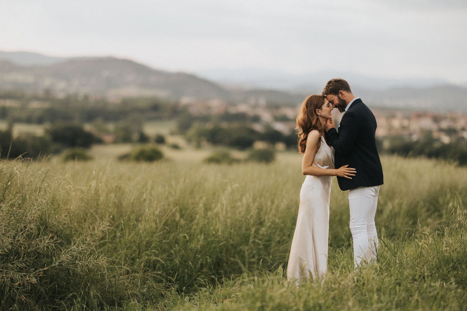 A couple stands together in an intimate embrace on a grassy Italian hillside with mountains in the distance.