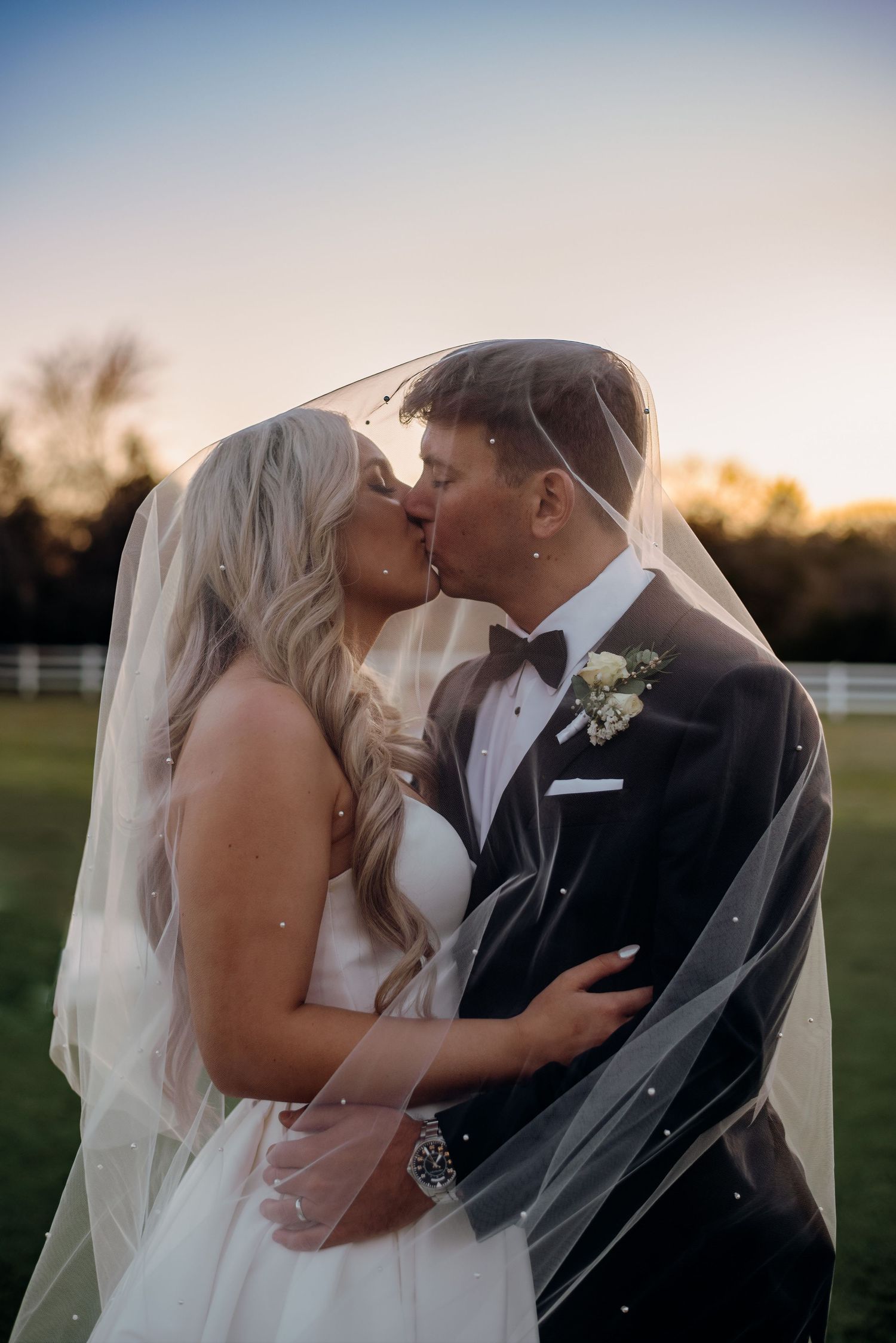 A romantic wedding kiss captured at dusk with a white fence and trees silhouetted in the background.