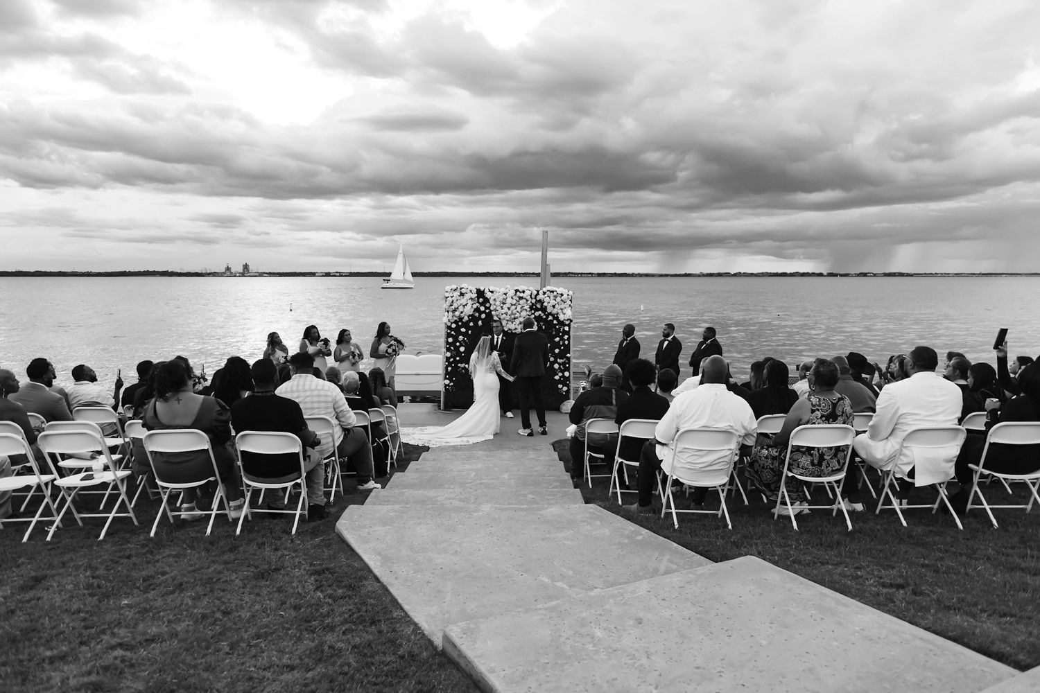 Black and white panoramic view of outdoor wedding ceremony setup with chairs facing water.