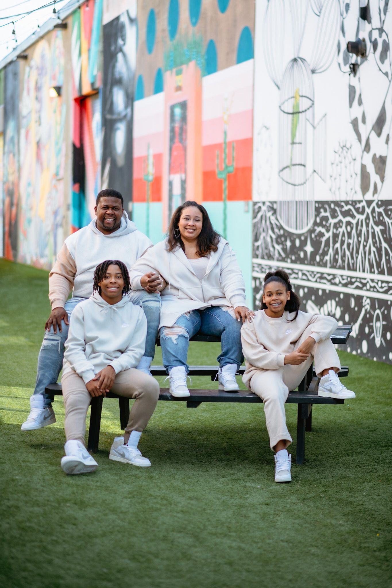A family sits together on a bench against a colorful mural wall, all wearing coordinated light-colored outfits.