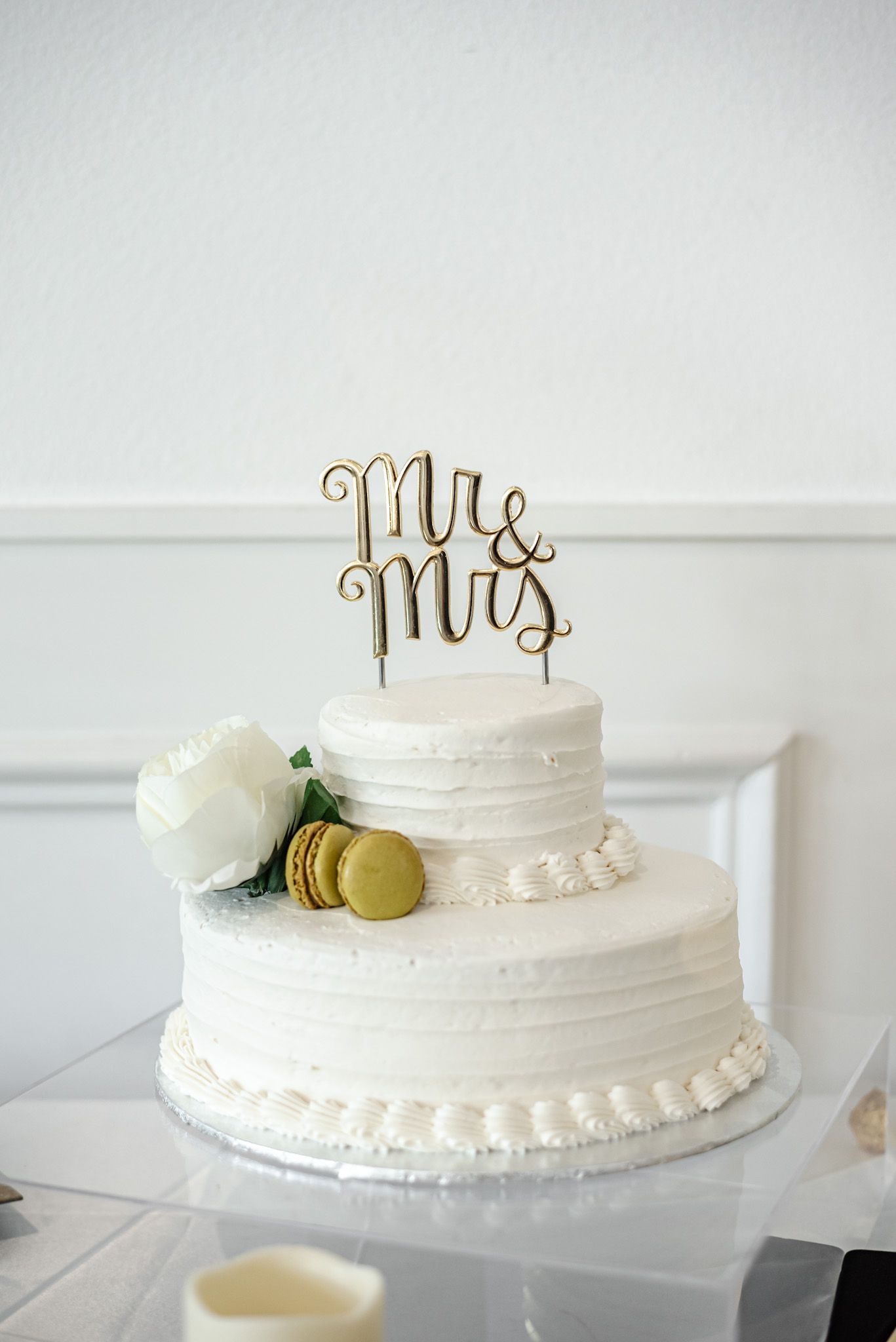Simple white wedding cake with gold 'Mr & Mrs' topper and minimal floral decoration.