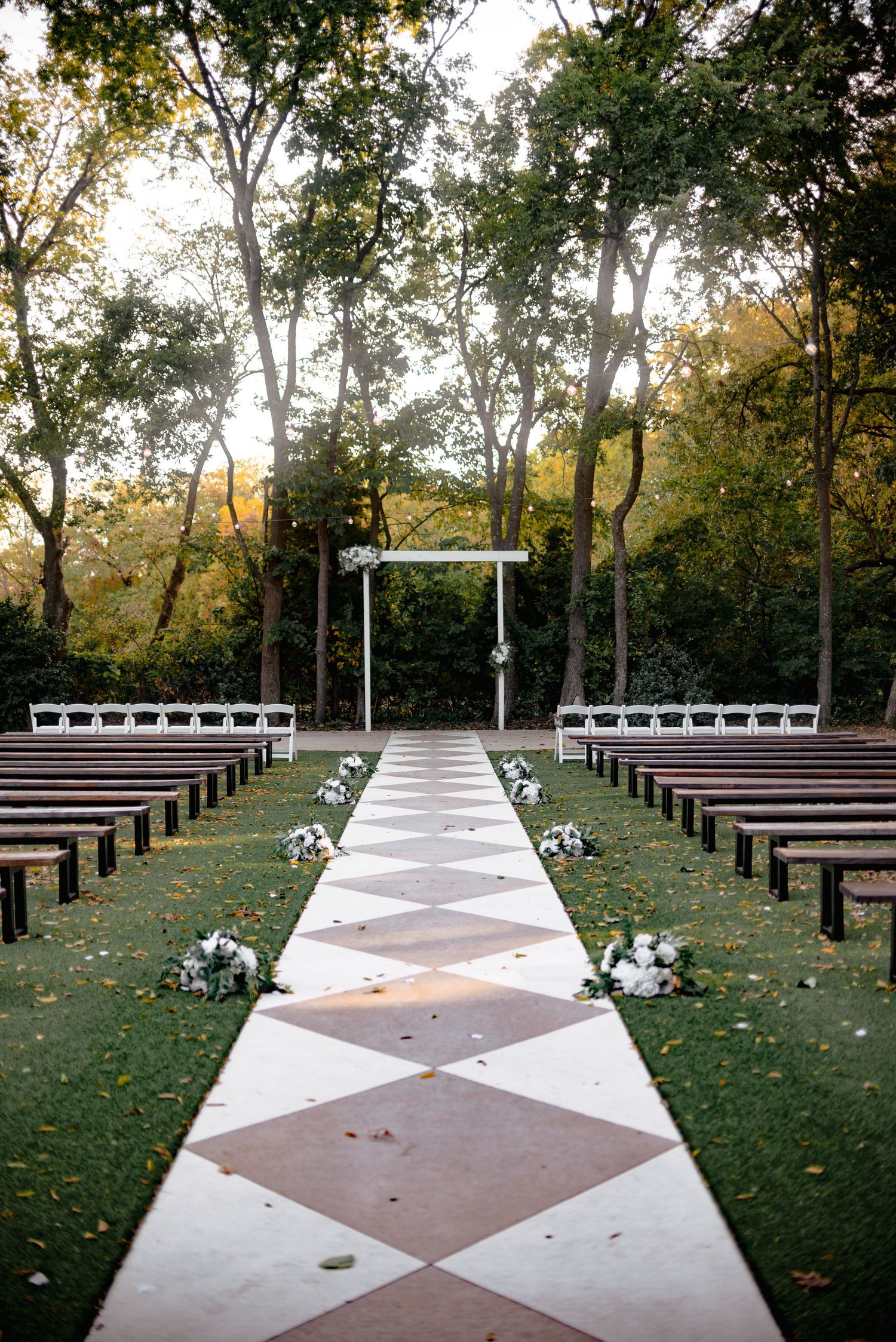 Outdoor wedding ceremony setup with wooden benches and white aisle runner surrounded by trees.