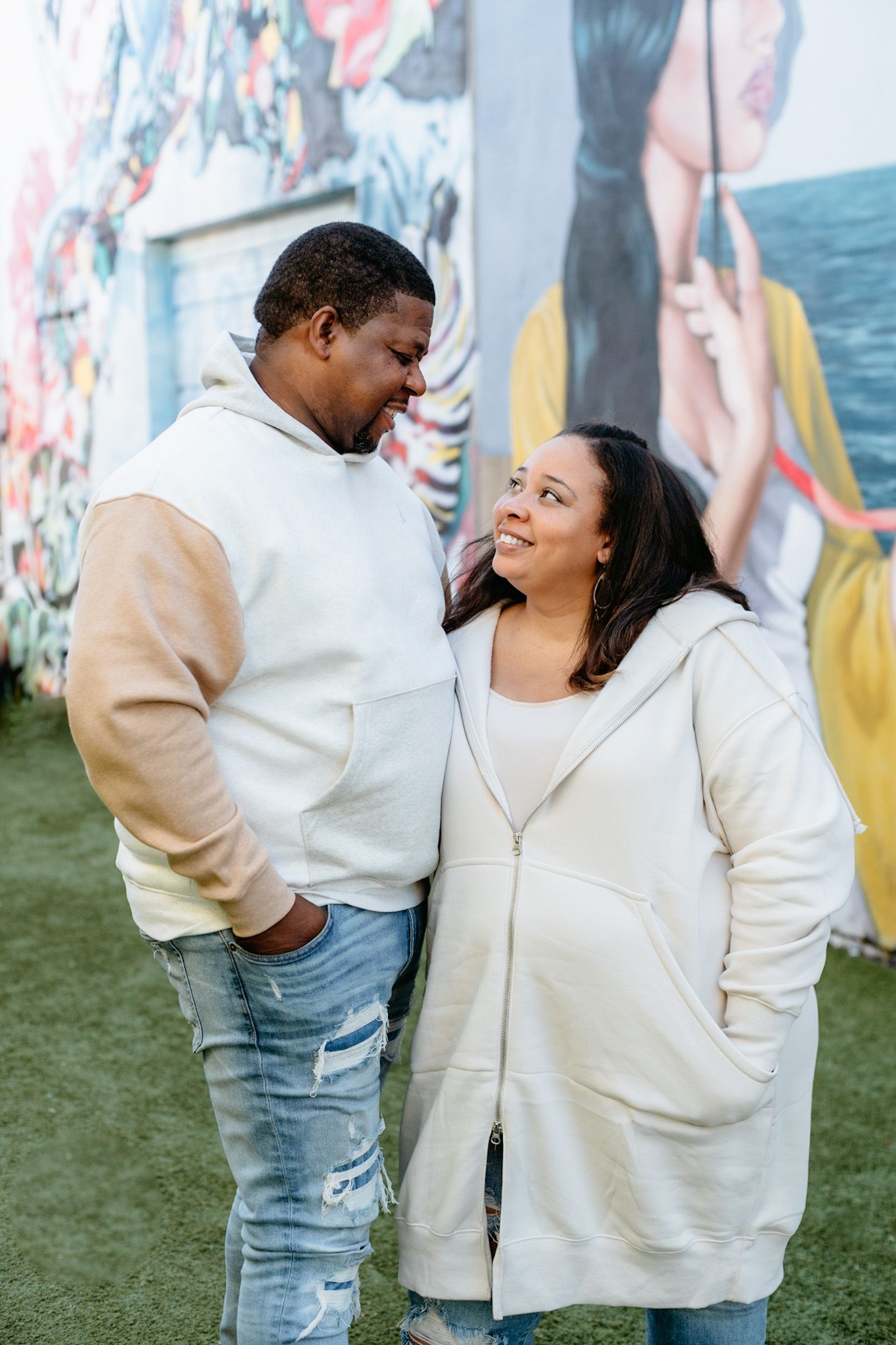 A couple in matching cream-colored hoodies share a tender moment against a mural backdrop.