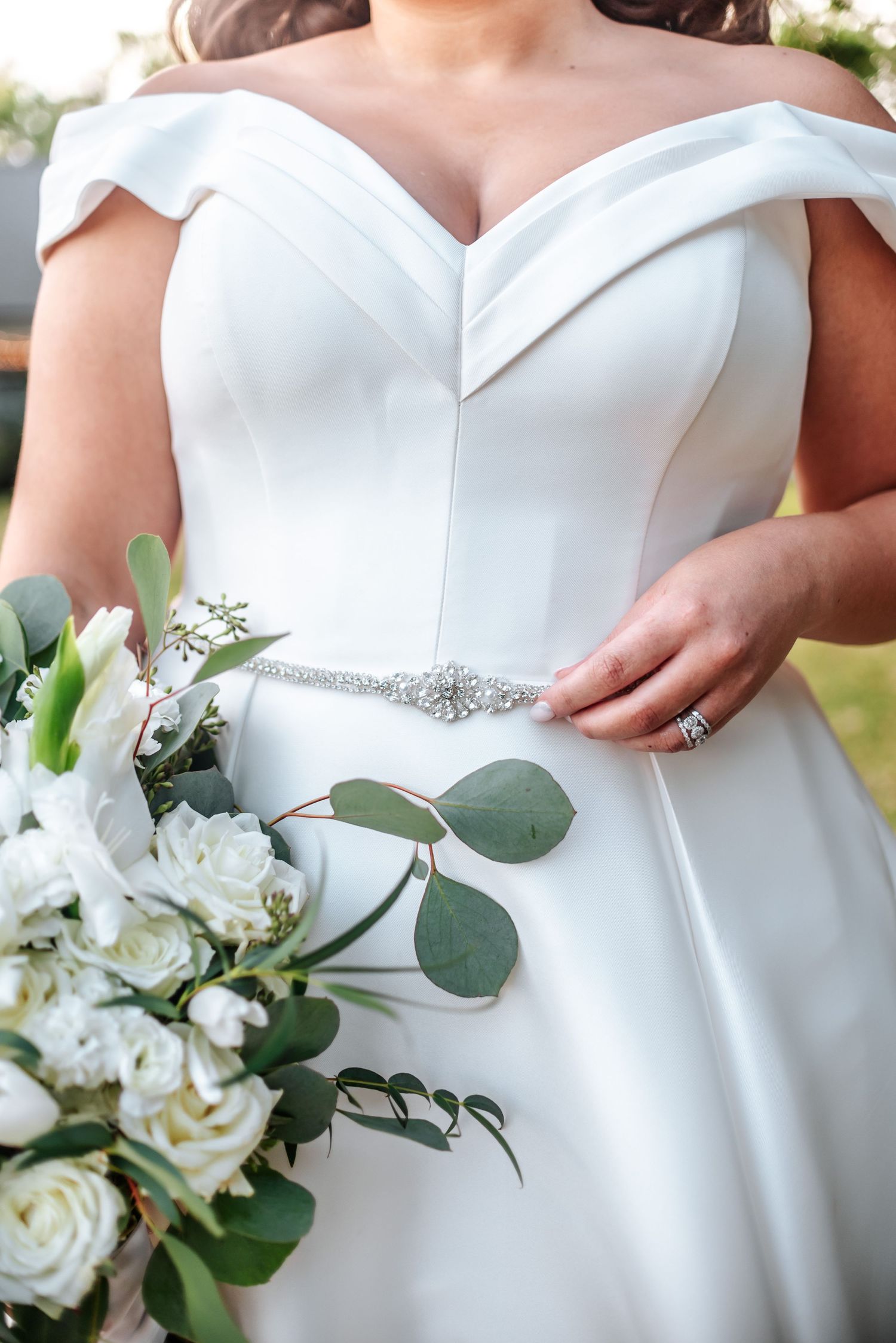 Detail shot of white wedding dress with off-shoulder neckline and floral bouquet.