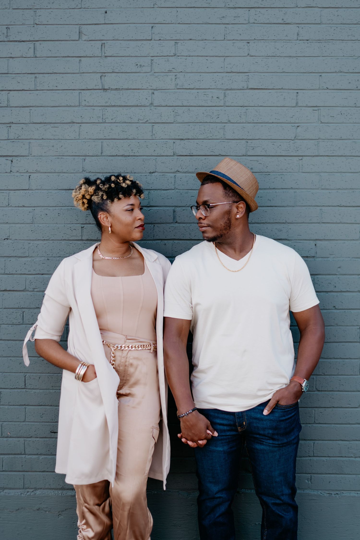 A stylish couple poses against a grey brick wall in neutral-toned outfits.