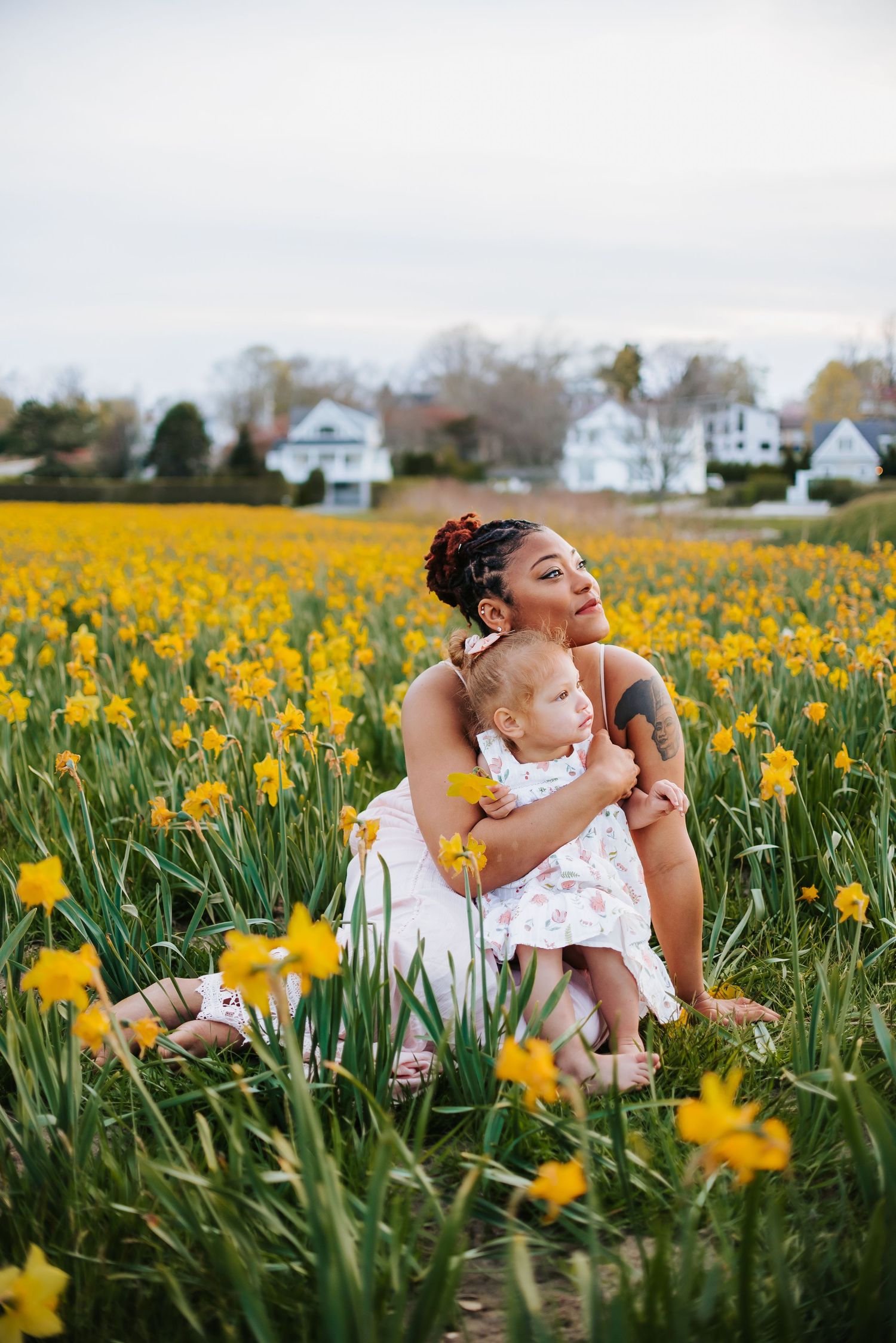 Mother and child laying in field of yellow daffodils with houses in background.
