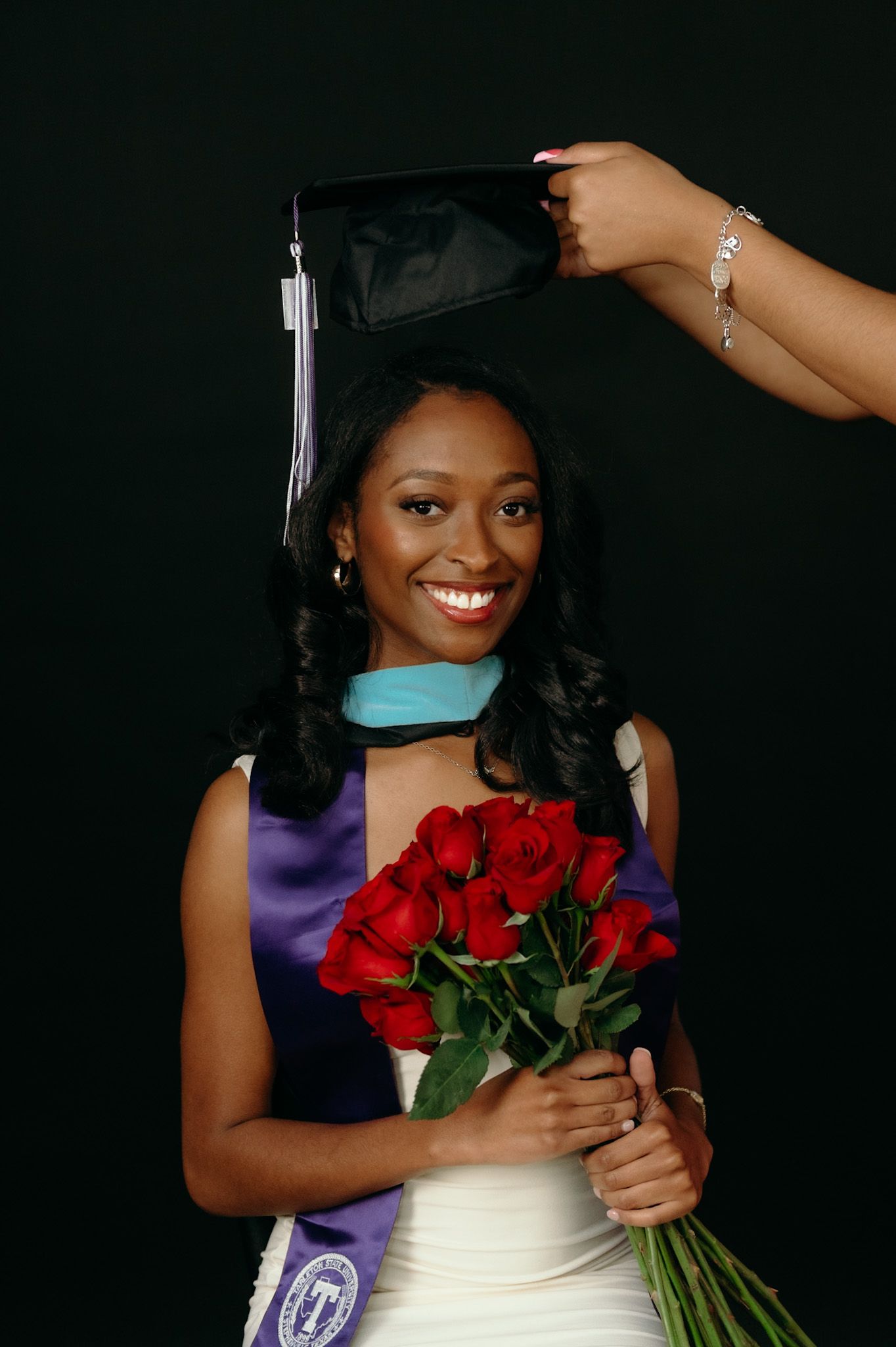 A graduate in academic regalia holds red roses while someone adjusts their graduation cap.