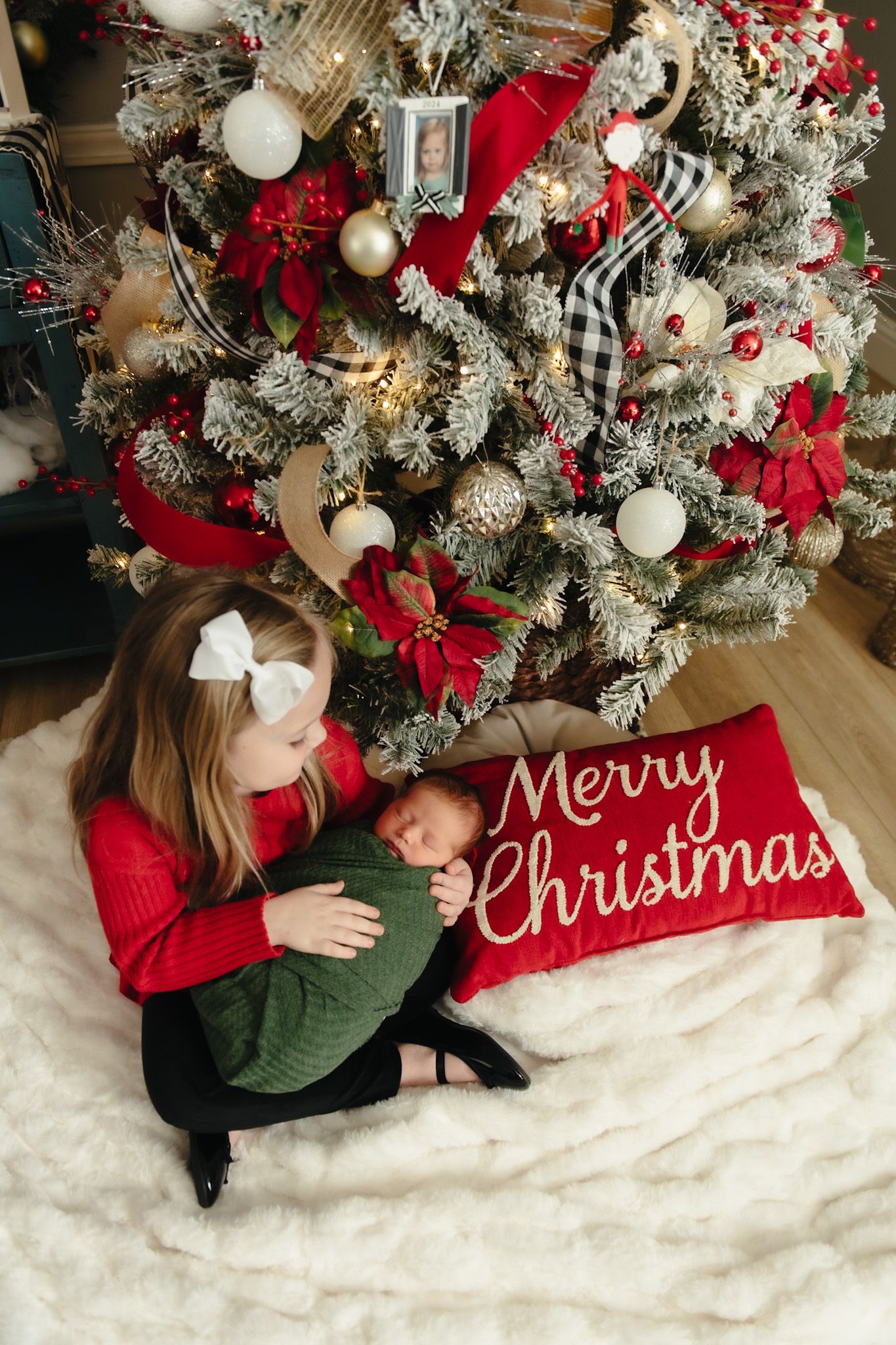 A decorated Christmas tree with a Merry Christmas pillow and child sitting beneath.
