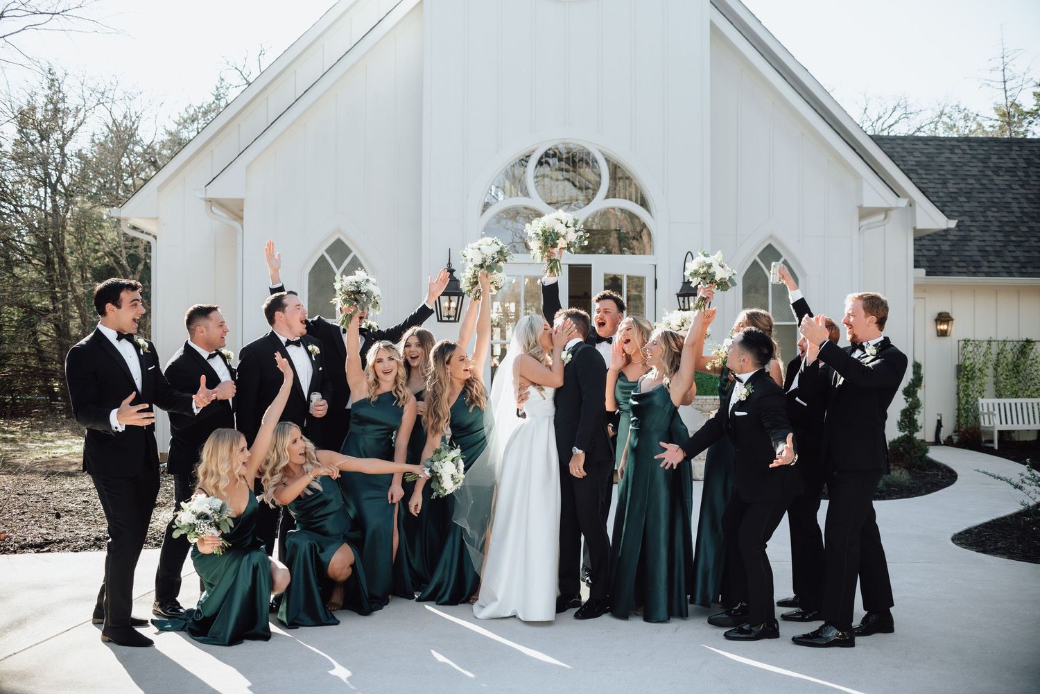 Wedding party in formal attire posing together outside white chapel.