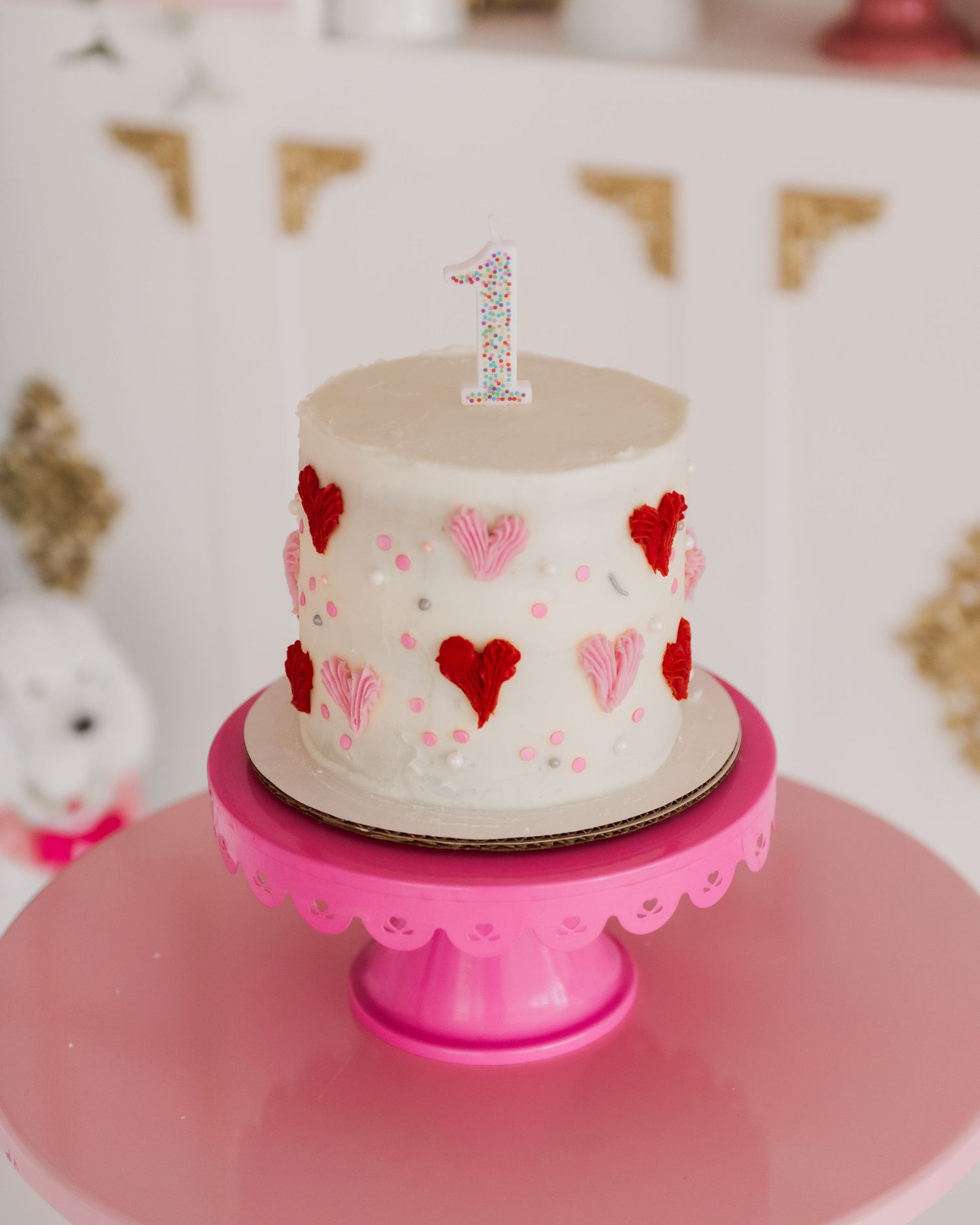 White birthday cake with pink hearts on pink cake stand against light background.