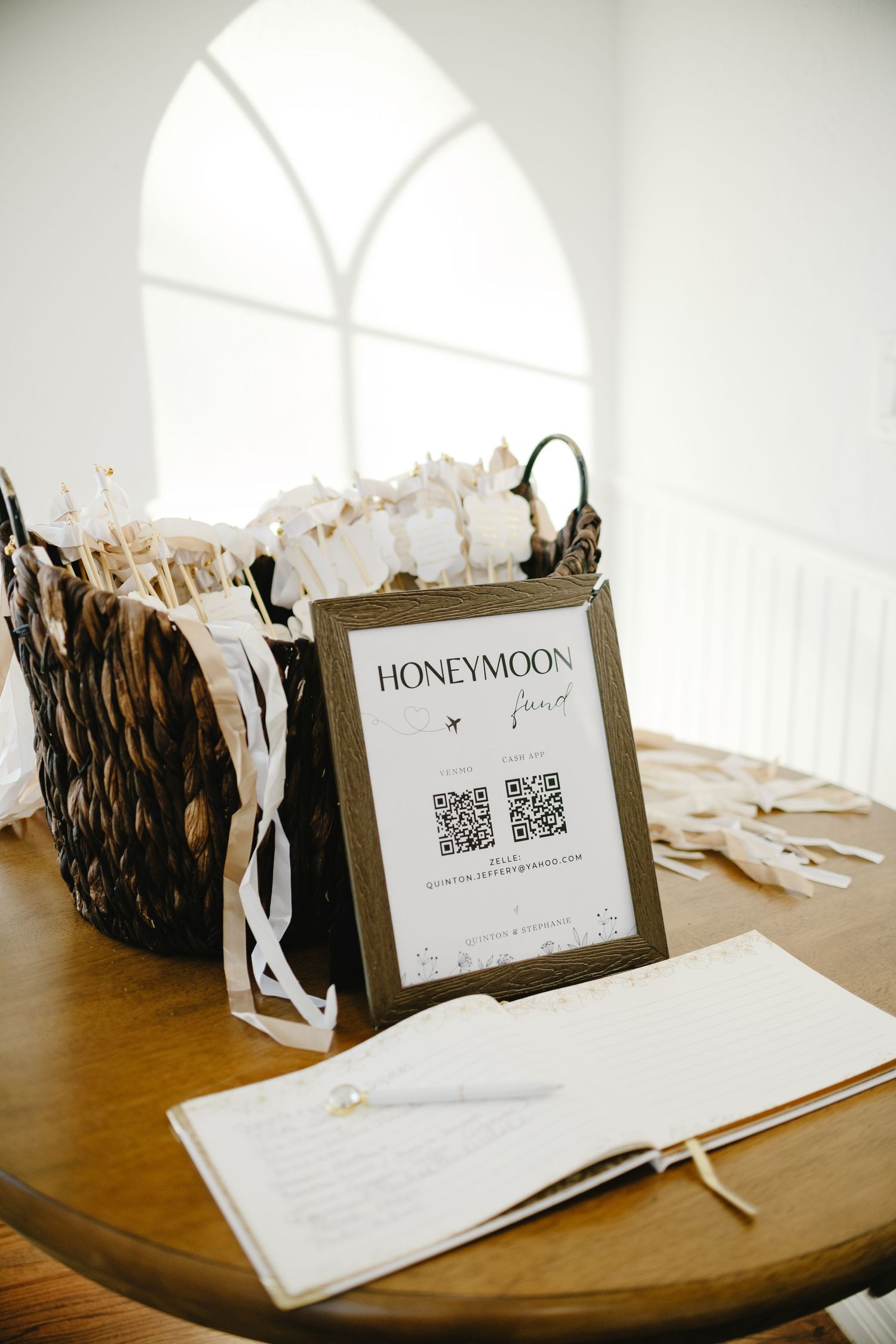 Wedding guestbook and Honeymoon Fund sign displayed on wooden table under arched windows.
