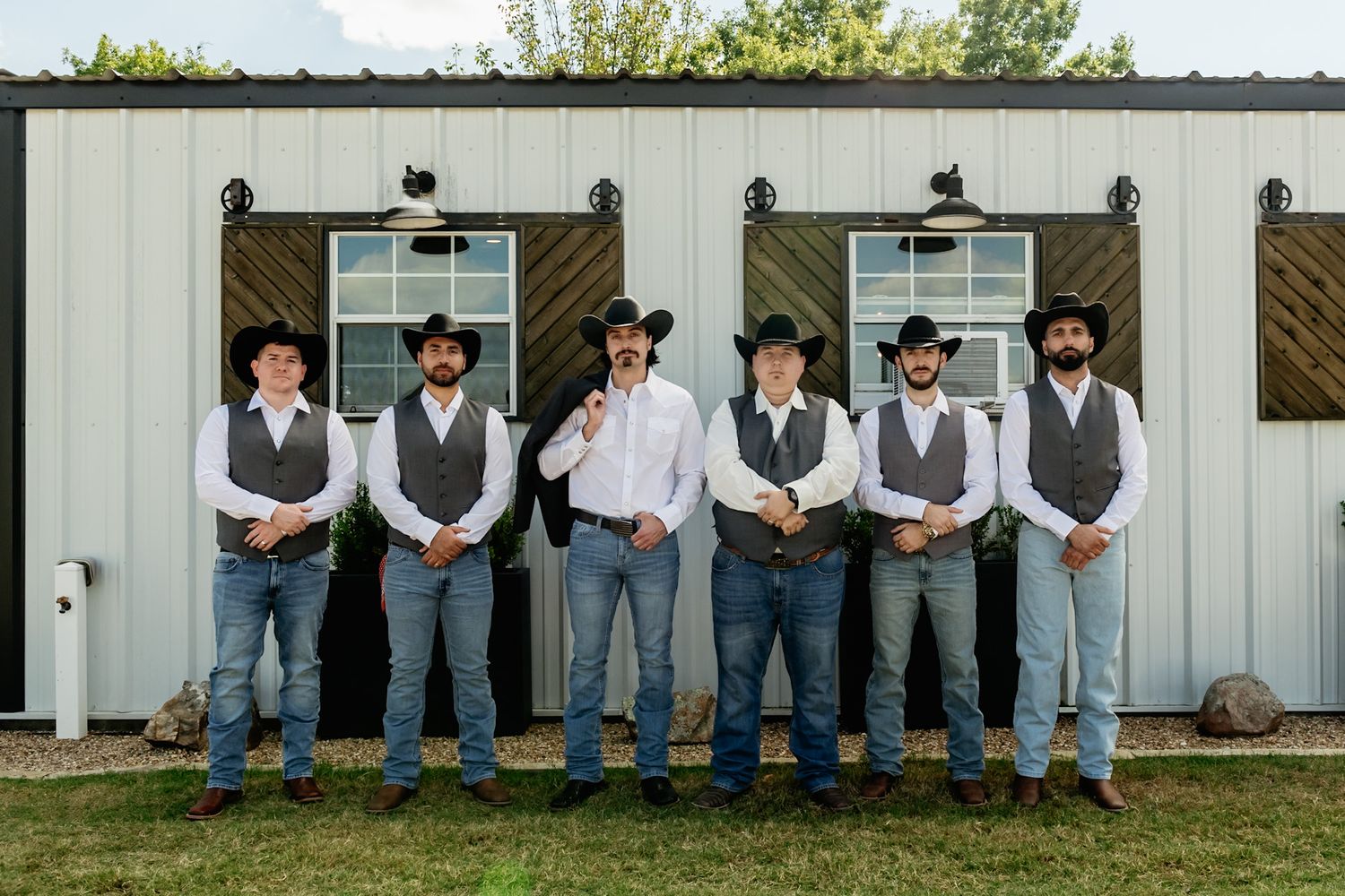 Groomsmen in matching vests and jeans lined up against white barn wall.