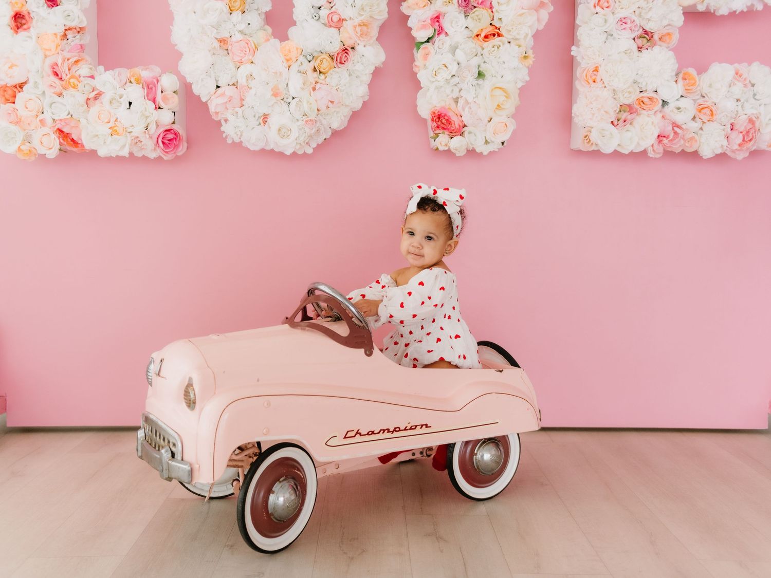 Pink vintage pedal car photographed in romantic floral studio setting.