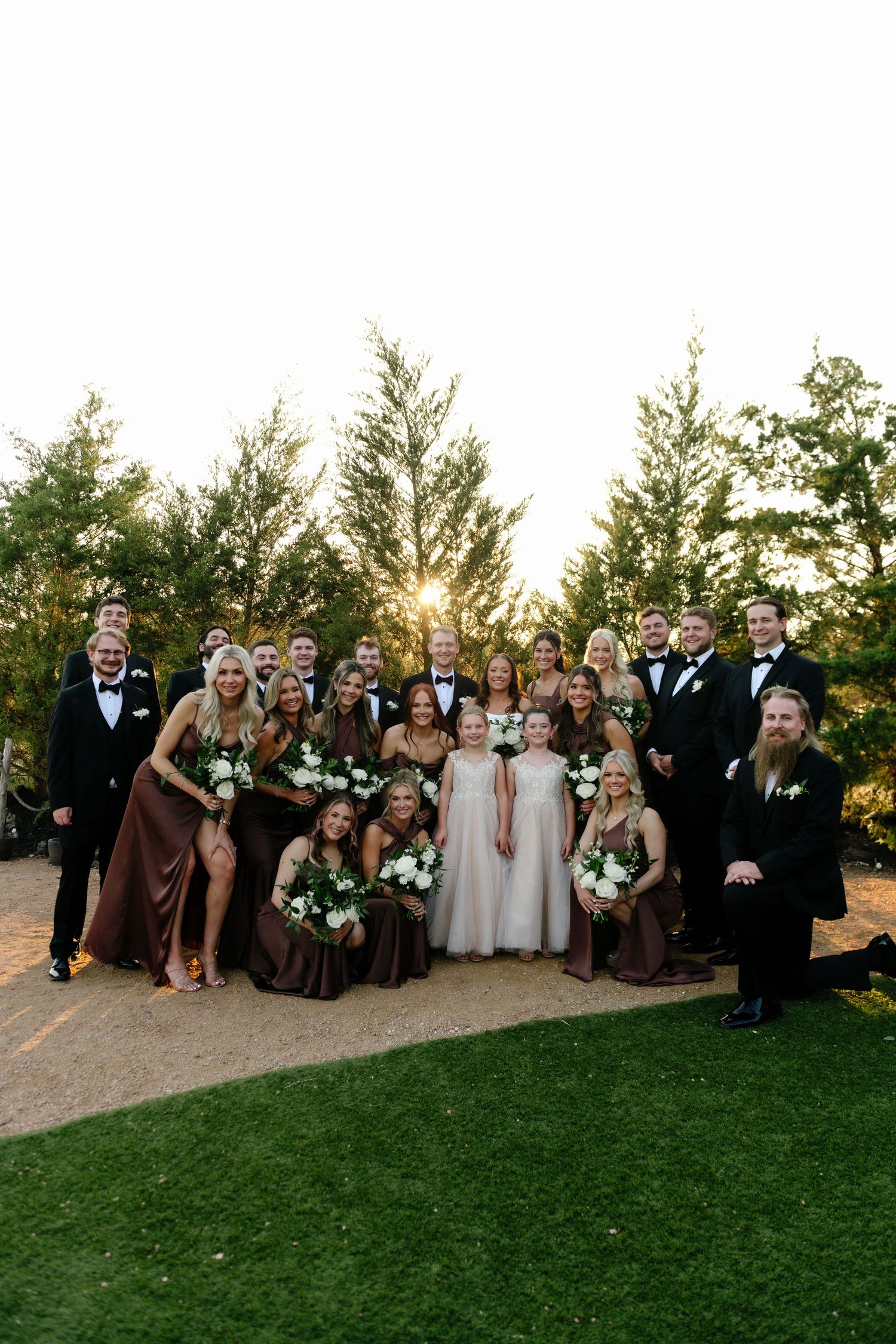 Wedding party gathered outdoors at sunset with trees in background.