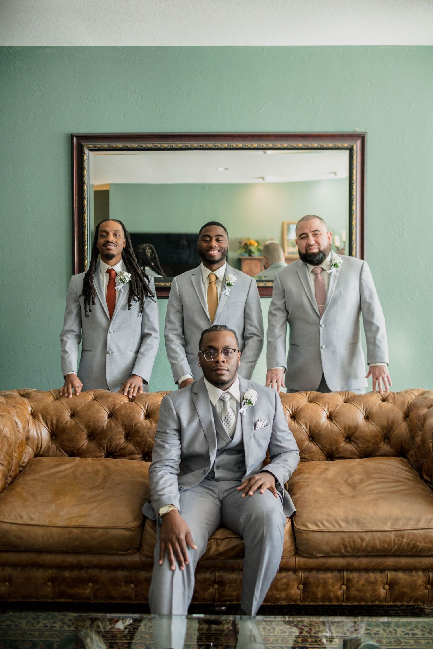 Groomsmen in light gray suits pose on a vintage brown leather couch in an elegant room.