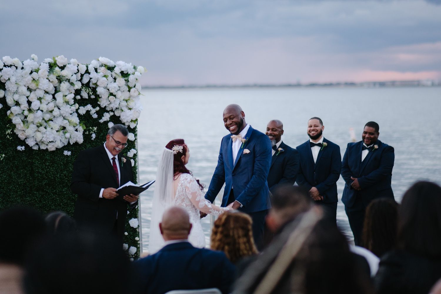 A sunset wedding ceremony by the water with a floral arch and guests watching the exchange of vows.