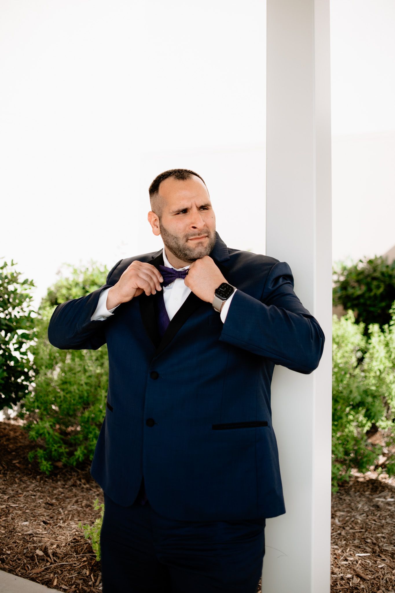 A person in a navy suit adjusts their bowtie while standing by a white column.