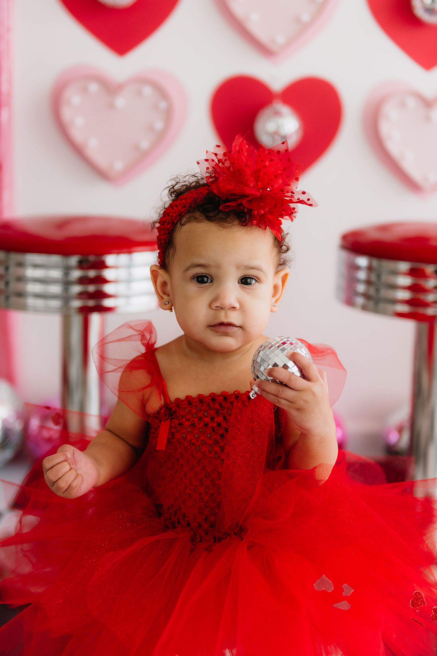 Baby in red tulle dress against pink and white heart-decorated background.