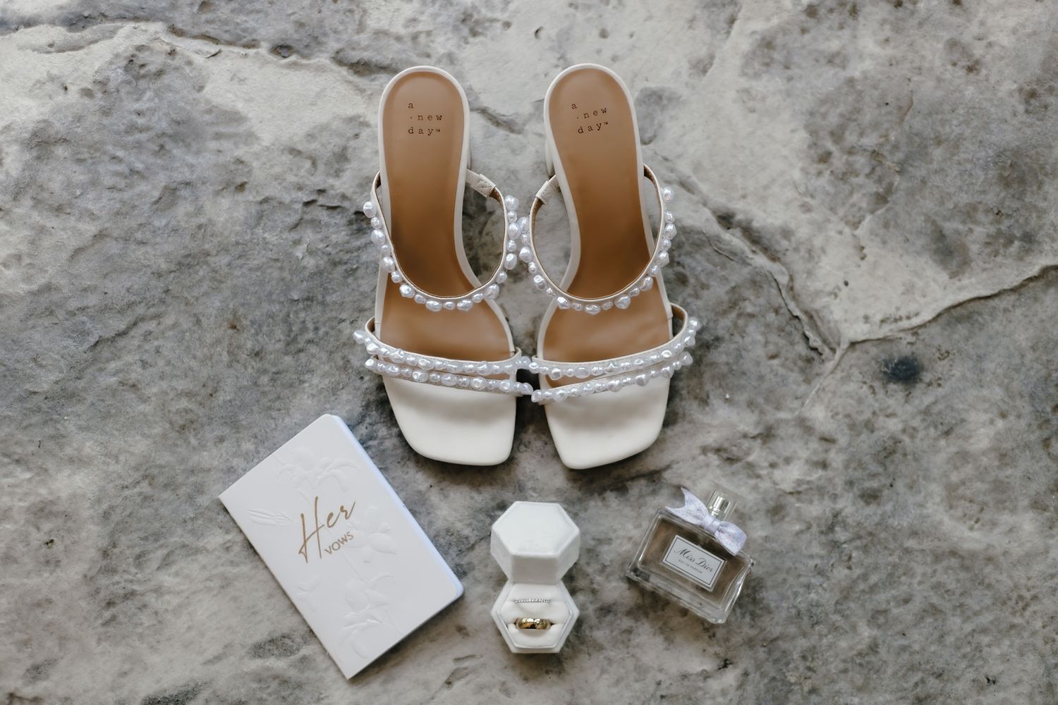 Wedding accessories including white heeled sandals, perfume bottle, ring box and vows card arranged on concrete surface.