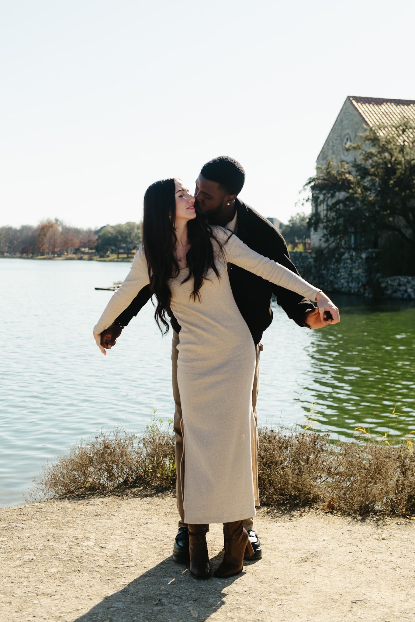 A playful couple poses by a lakeside with a stone building in the distance.