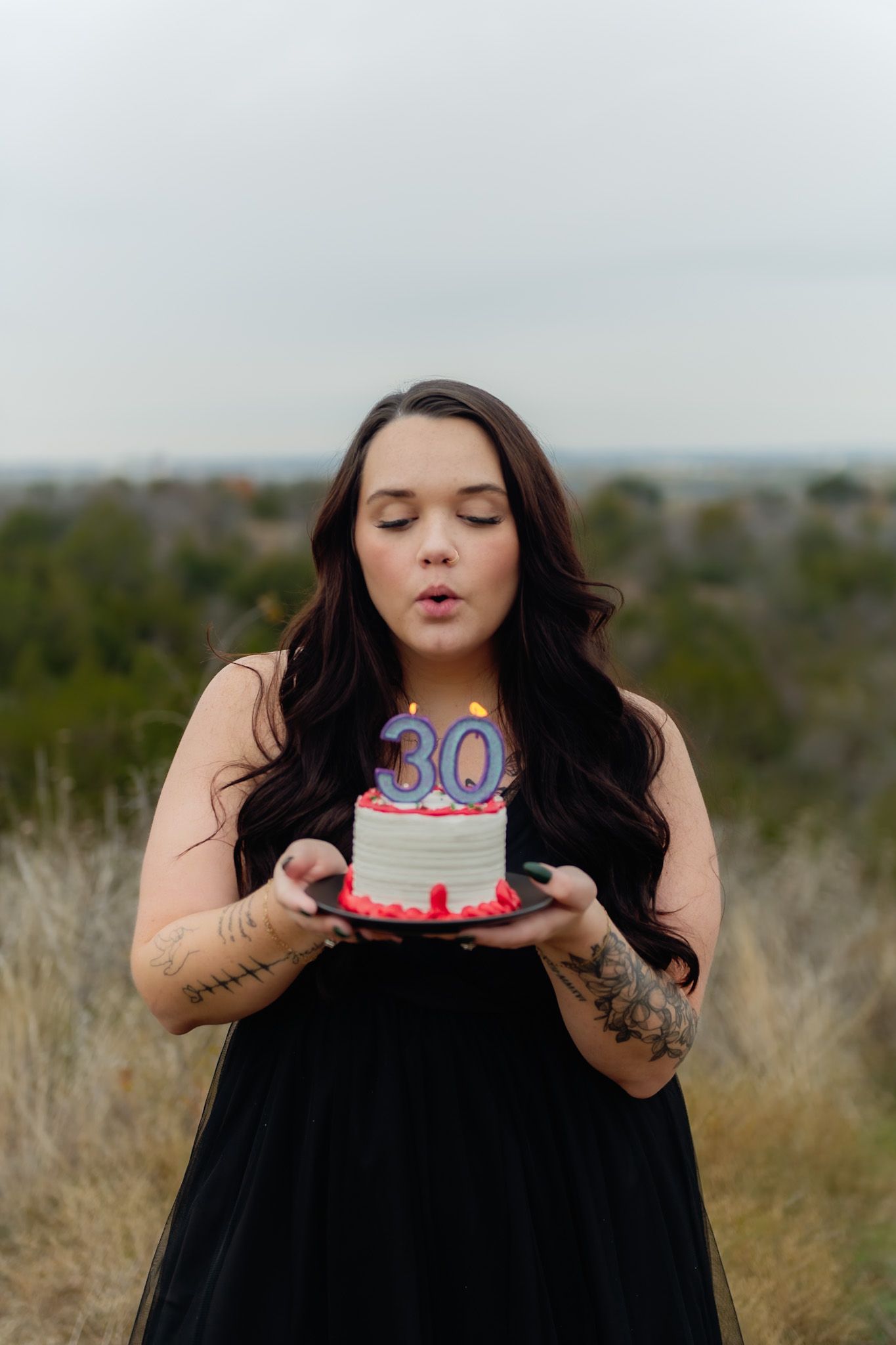 Person in black dress holding birthday cake outdoors in grassy field.