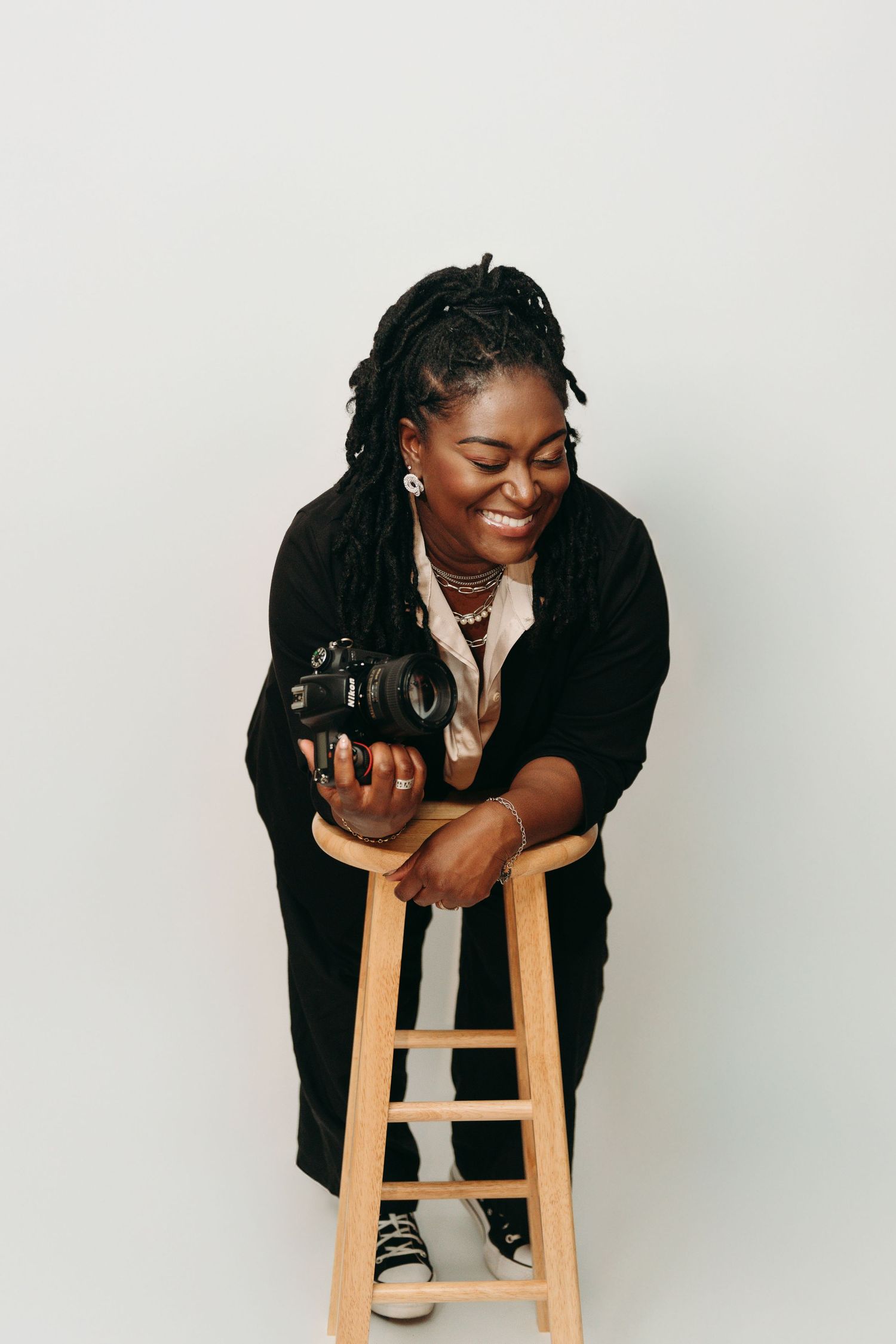 Professional photographer smiling while leaning on wooden stool with camera in studio setting.