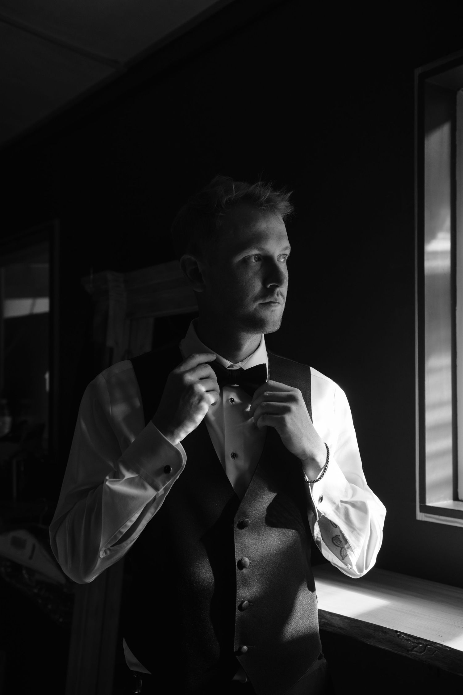 Dramatic black and white portrait of groom adjusting bowtie near window.